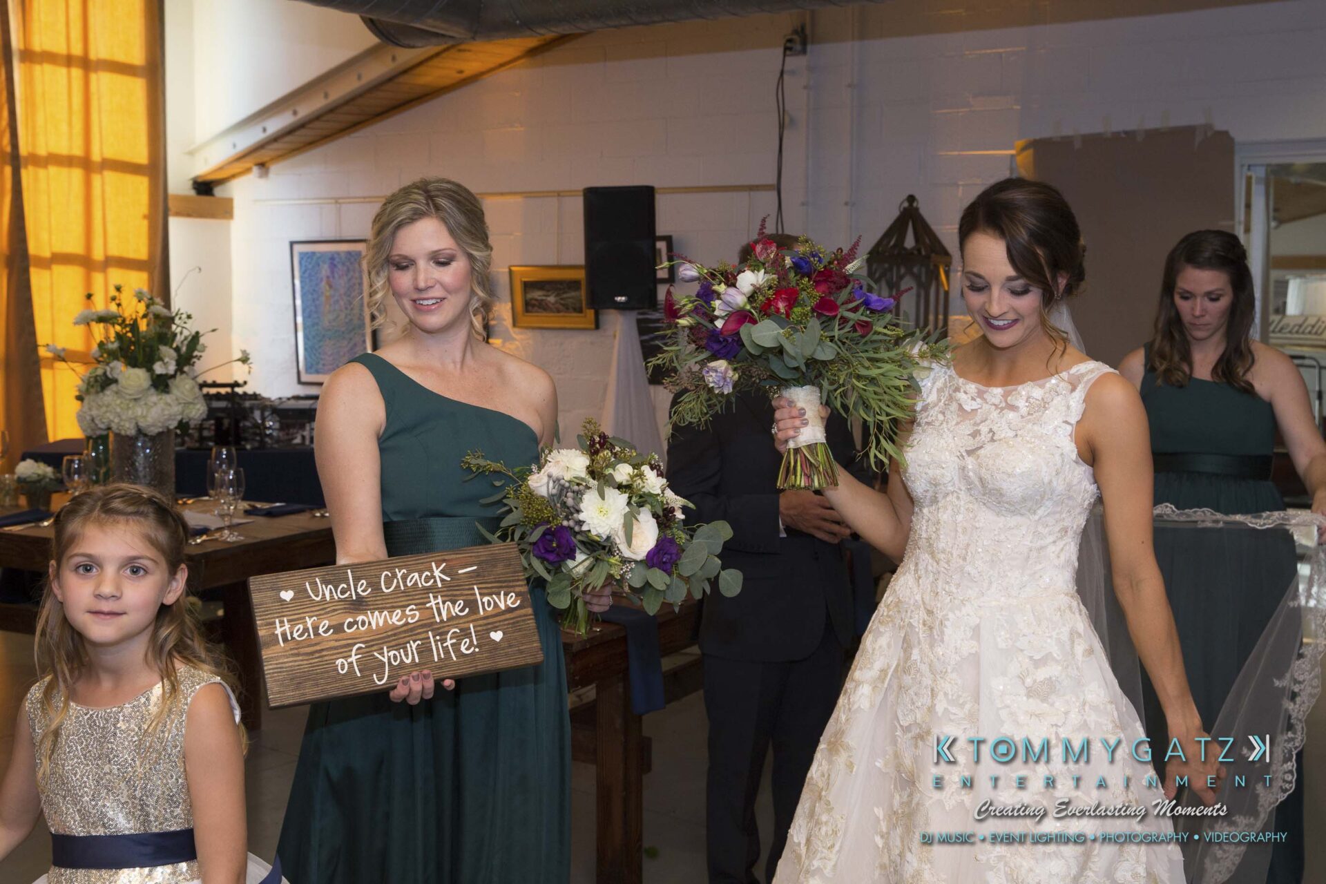 Amanda-Tyler-102117-CEREMONY-Low-Res-WM-3 - TOMMY GATZ ENTERTAINMENT Bridesmaid holds a sign saying "Uncle Crack — Here comes the love of your life!" while bride in a white dress carries a bouquet, standing with a young girl and other bridesmaids.