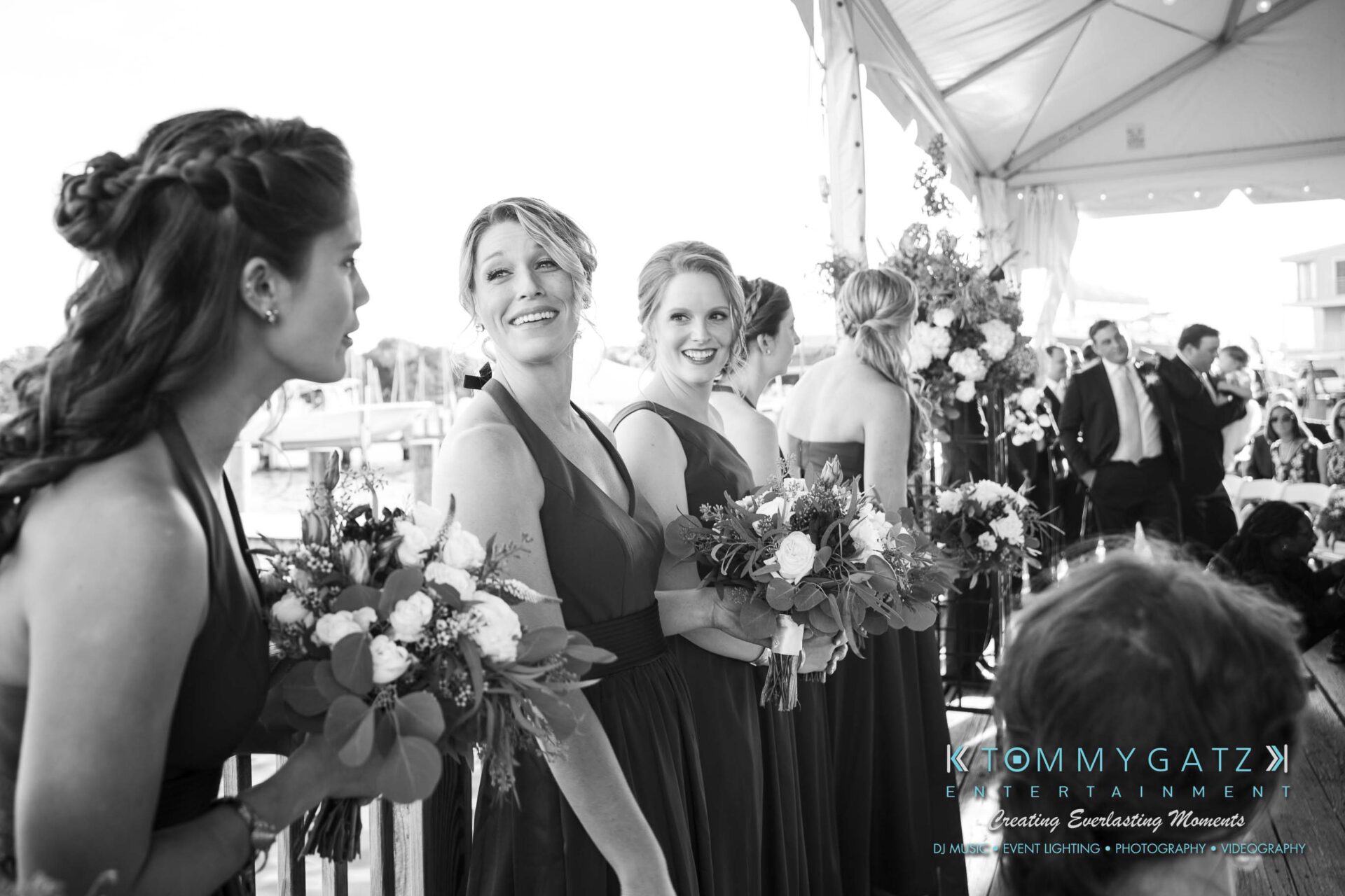 Amanda-Tyler-102117-CEREMONY-Low-Res-WM-30 - TOMMY GATZ ENTERTAINMENT Bridesmaids in dark dresses stand holding bouquets, smiling under a tent at an outdoor event.