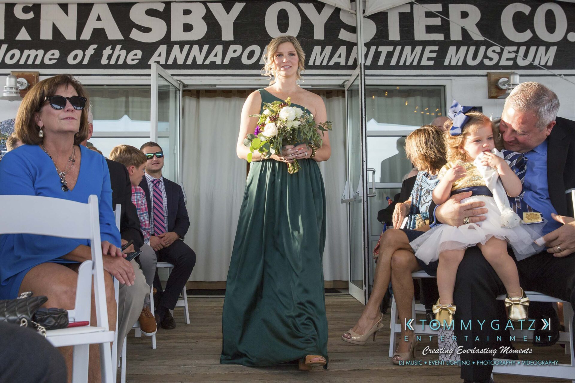 Amanda-Tyler-102117-CEREMONY-Low-Res-WM-31 - TOMMY GATZ ENTERTAINMENT A woman in a green dress walks down an aisle at an outdoor event, carrying a bouquet. Guests are seated on either side, and a man is holding a young child on the right.