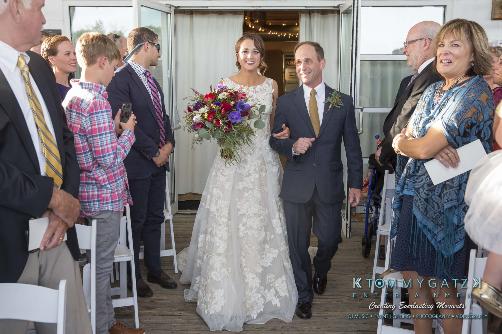 Amanda-Tyler-102117-CEREMONY-Low-Res-WM-36 - TOMMY GATZ ENTERTAINMENT Bride in a white gown walks down the aisle with a man, holding a vibrant bouquet, surrounded by smiling guests in outdoor setting.
