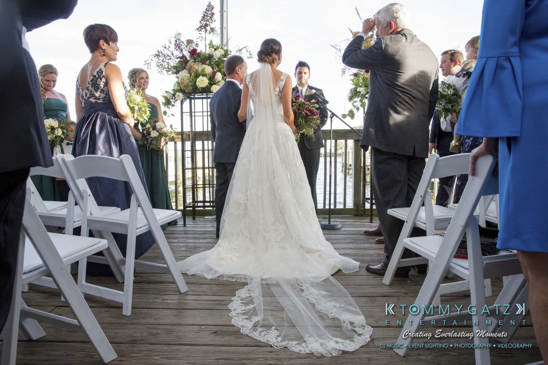Amanda-Tyler-102117-CEREMONY-Low-Res-WM-38 - TOMMY GATZ ENTERTAINMENT Bride in a white dress walks down the aisle on a wooden deck, with guests seated on either side and floral arrangements in the background.