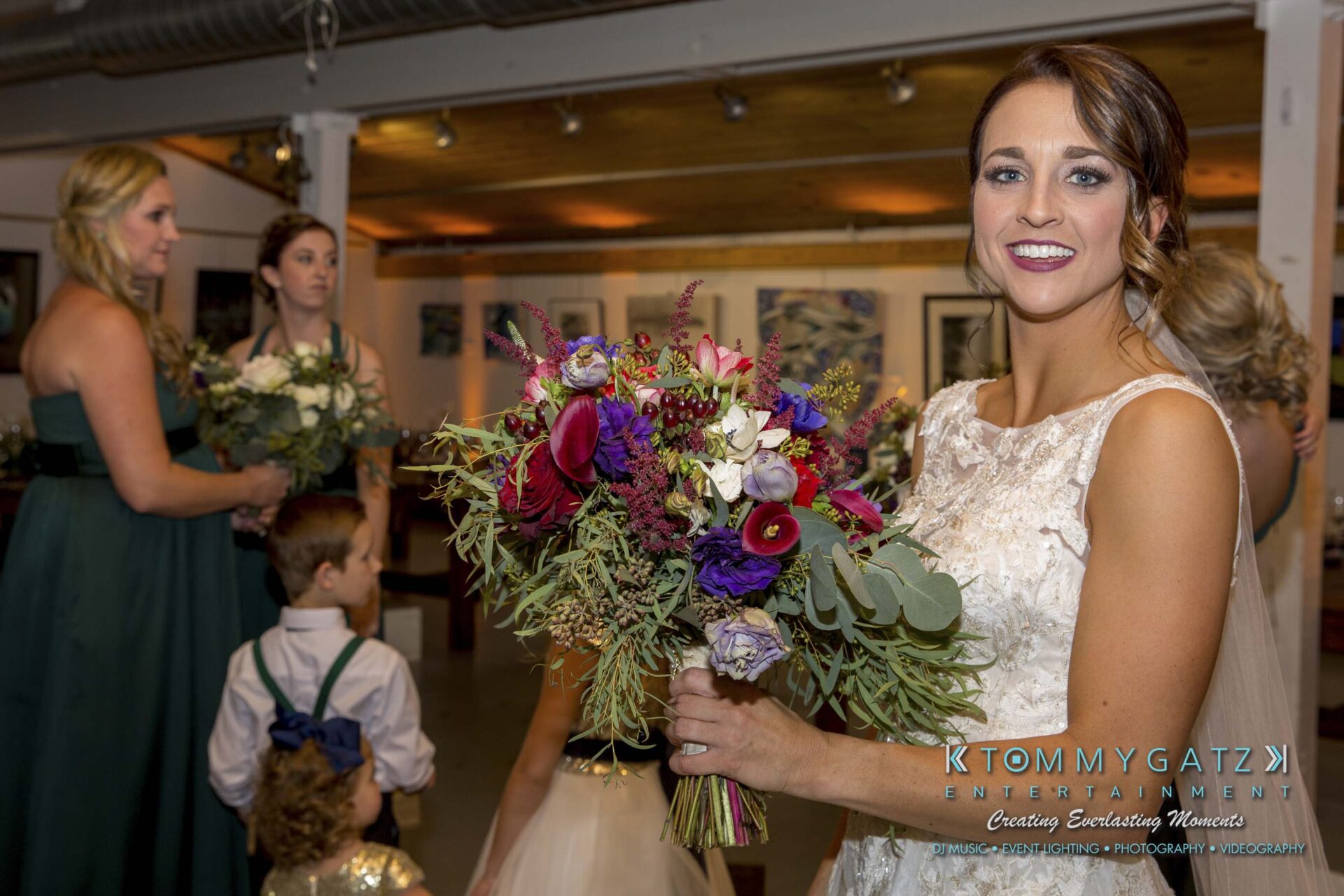 A bride holds a colorful bouquet, smiling at the camera. Bridesmaids and children are in the background in an indoor setting.