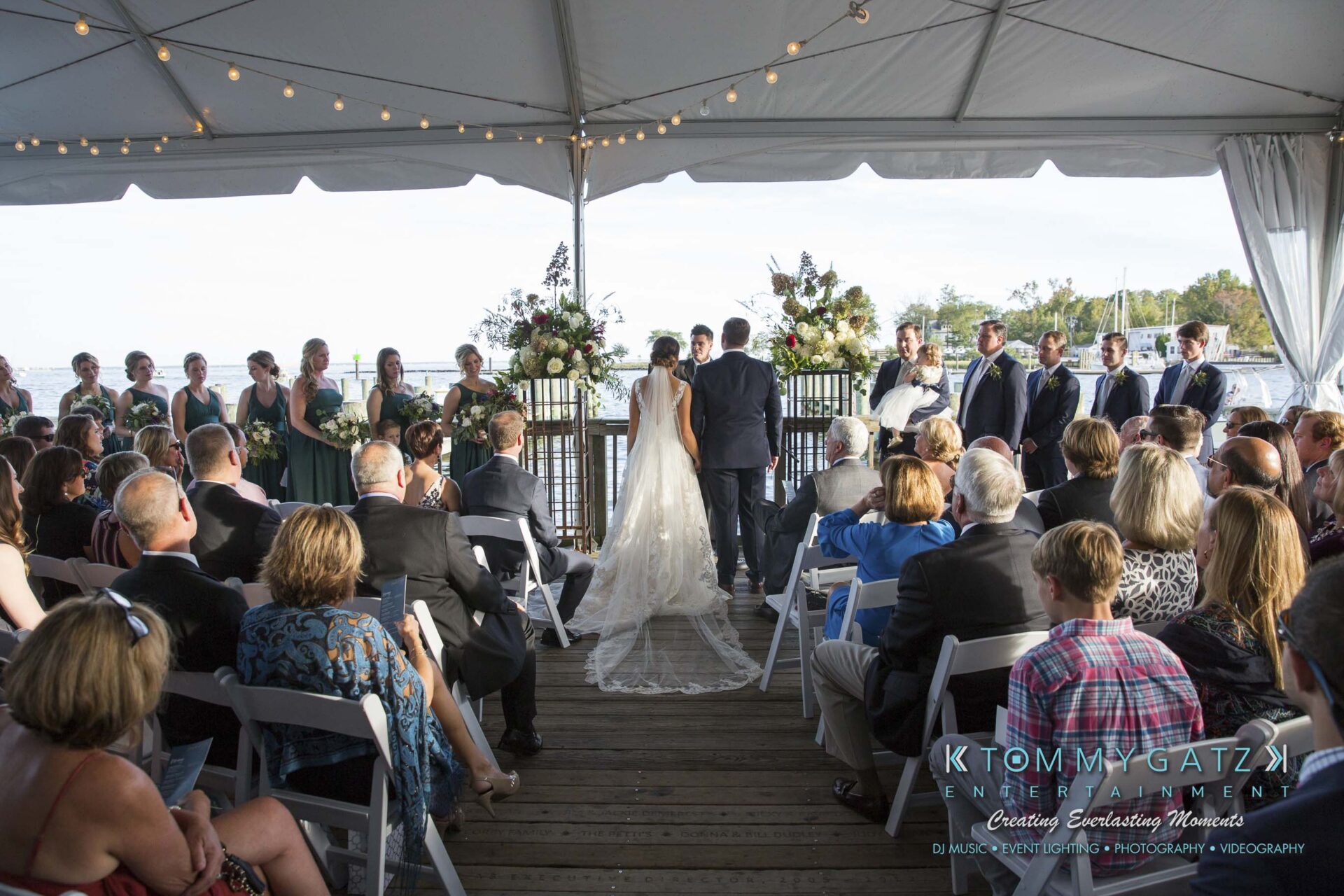 Amanda-Tyler-102117-CEREMONY-Low-Res-WM-46 - TOMMY GATZ ENTERTAINMENT A wedding ceremony under a tent by the water. The couple stands at the altar surrounded by the bridal party and guests seated on white chairs.