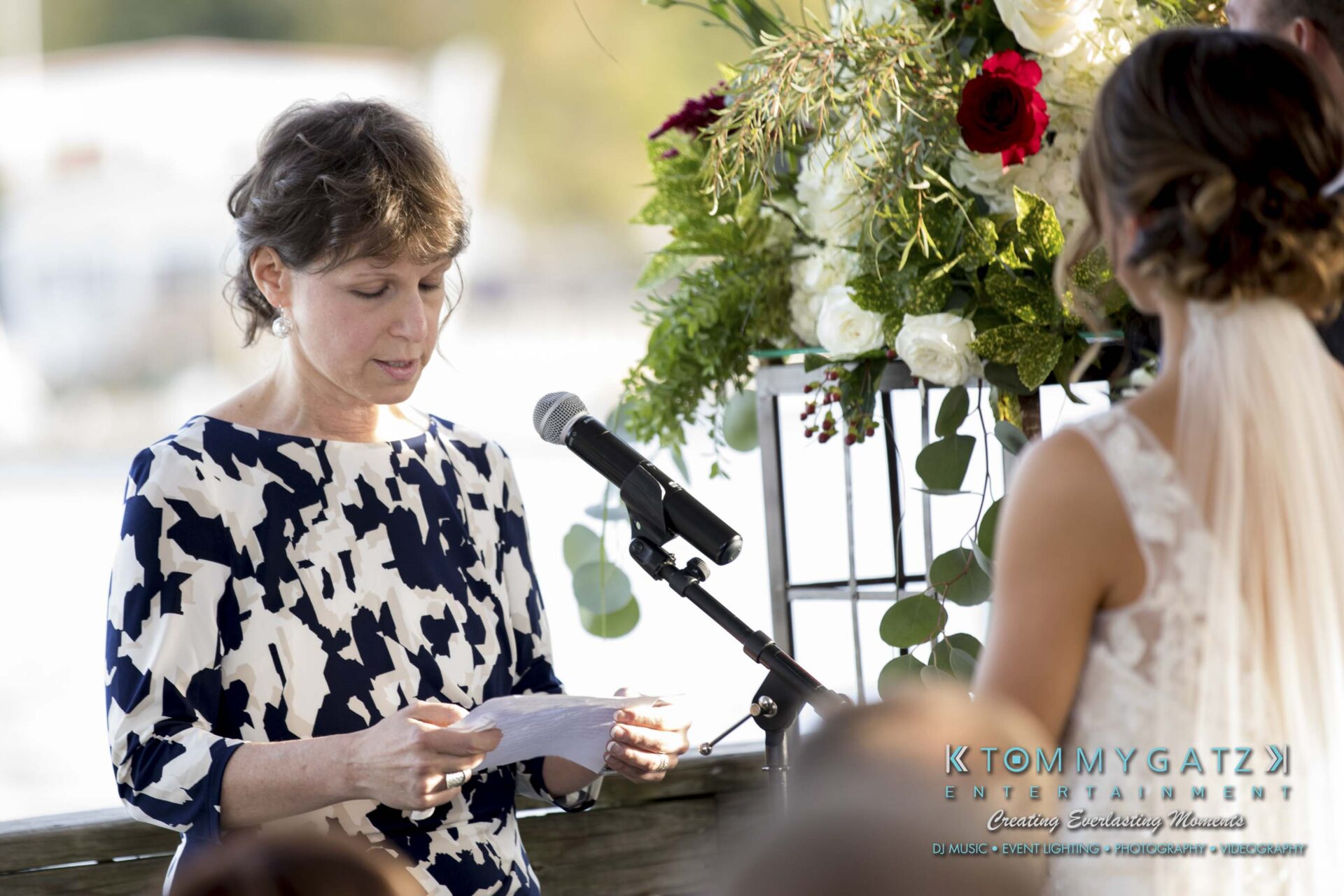 A woman in a patterned dress speaks into a microphone while holding a piece of paper. Another person, with their back to the camera, listens. A floral arrangement is visible in the background.