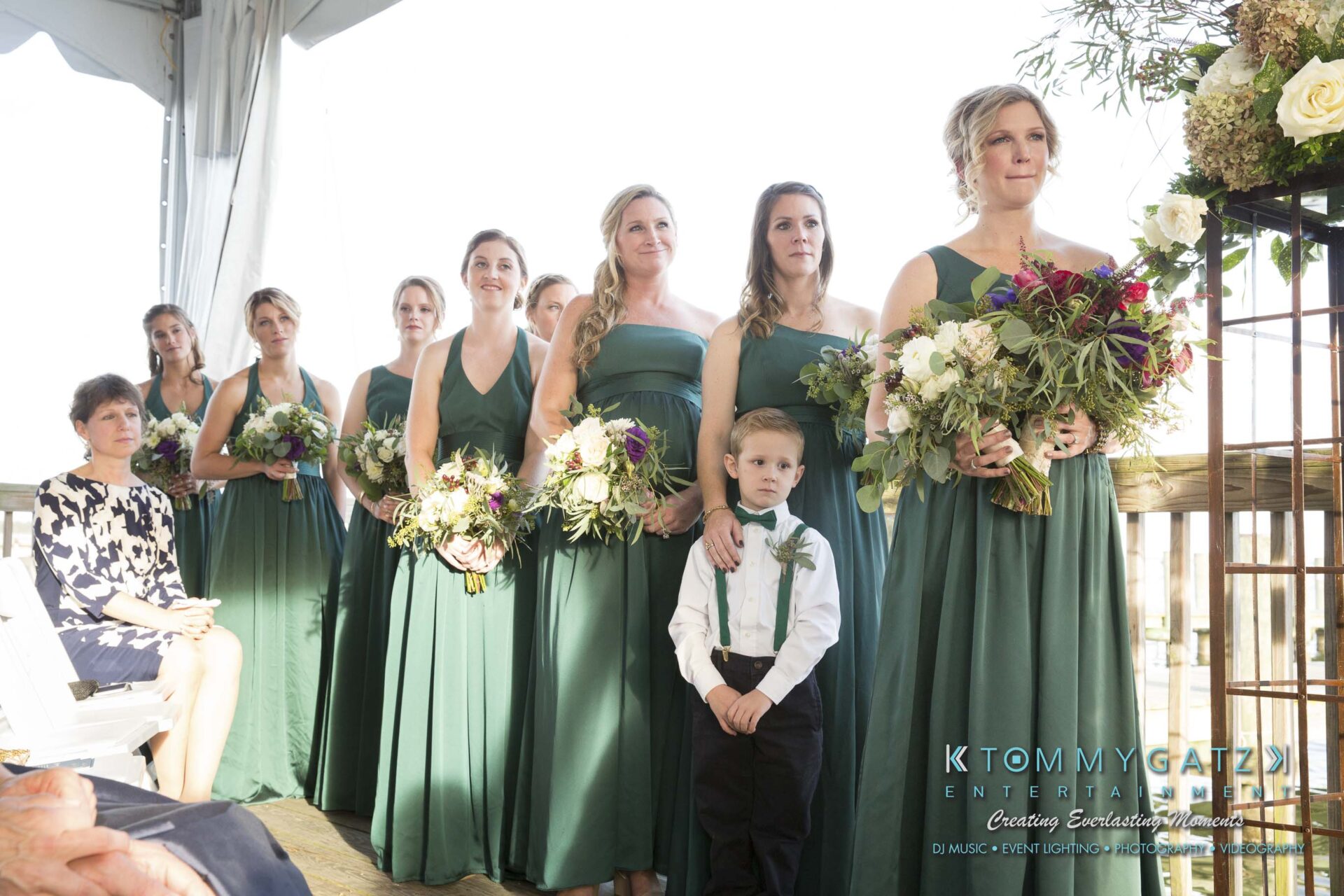 Amanda-Tyler-102117-CEREMONY-Low-Res-WM-56 - TOMMY GATZ ENTERTAINMENT A group of bridesmaids in green dresses stand with bouquets. A young boy in formal attire stands in front. People are seated nearby.