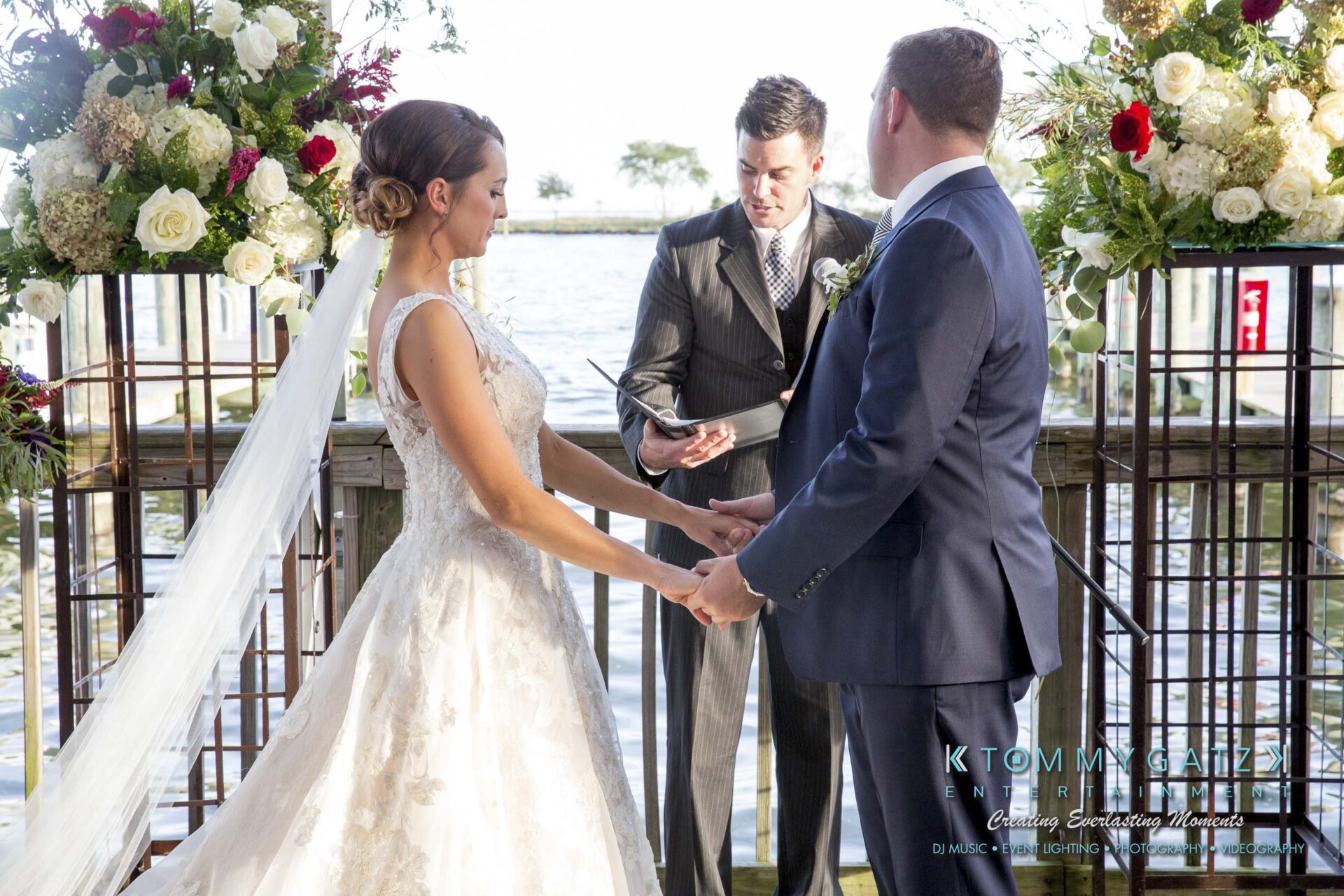 Amanda-Tyler-102117-CEREMONY-Low-Res-WM-59 - TOMMY GATZ ENTERTAINMENT A couple holds hands during a wedding ceremony by the water, with a person officiating. Floral arrangements frame the scene.