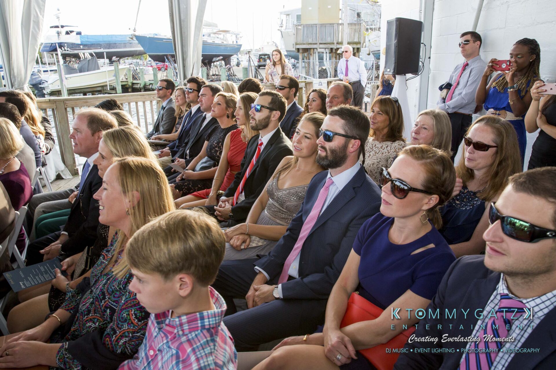 Amanda-Tyler-102117-CEREMONY-Low-Res-WM-62 - TOMMY GATZ ENTERTAINMENT People seated outdoors at an event near a marina, with boats in the background. Some attendees wear sunglasses, and there's a mix of formal and casual attire.