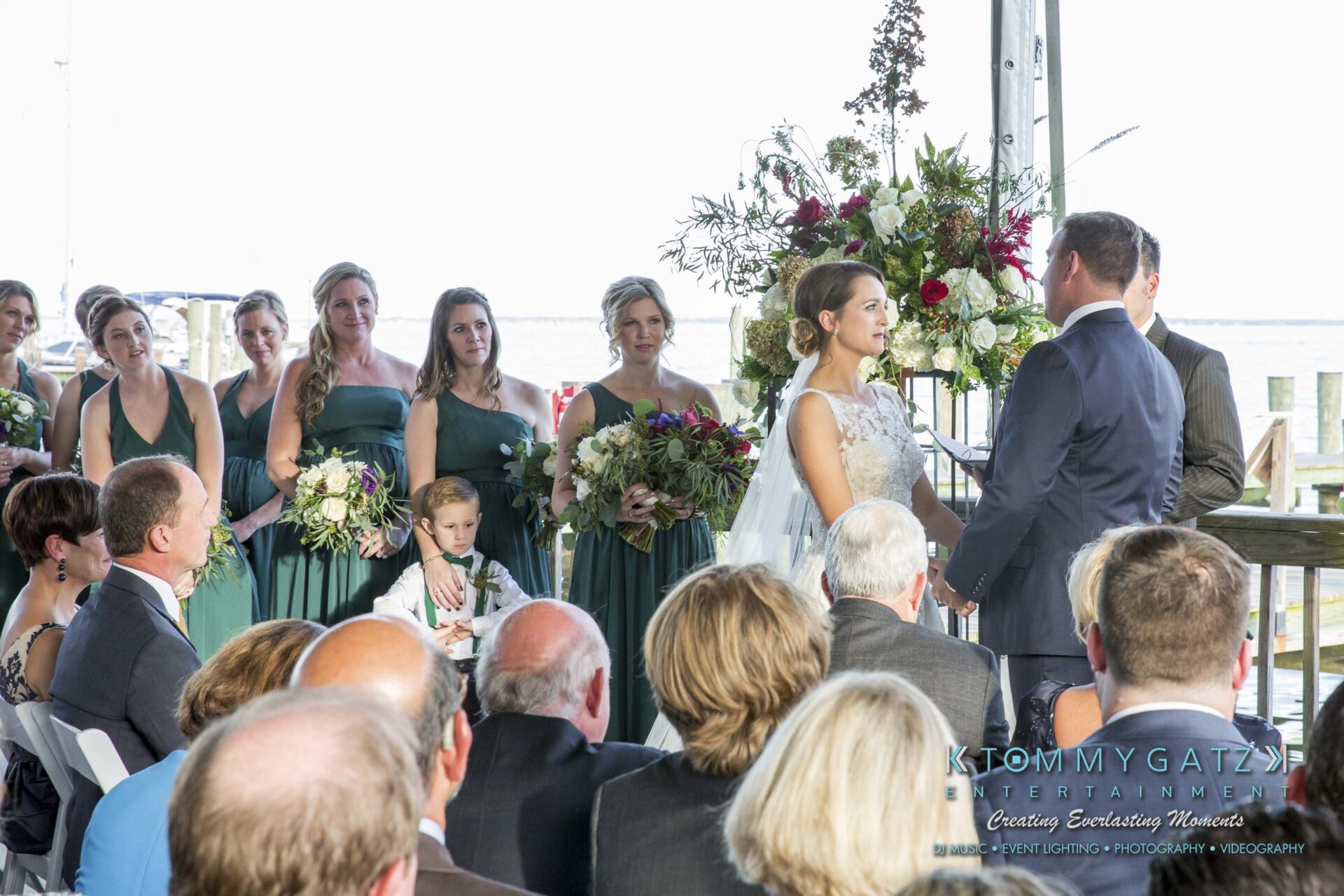 Amanda-Tyler-102117-CEREMONY-Low-Res-WM-64 - TOMMY GATZ ENTERTAINMENT A couple exchange vows at an outdoor wedding ceremony, with bridesmaids in green dresses and a flower girl in attendance. Guests are seated, observing the event.