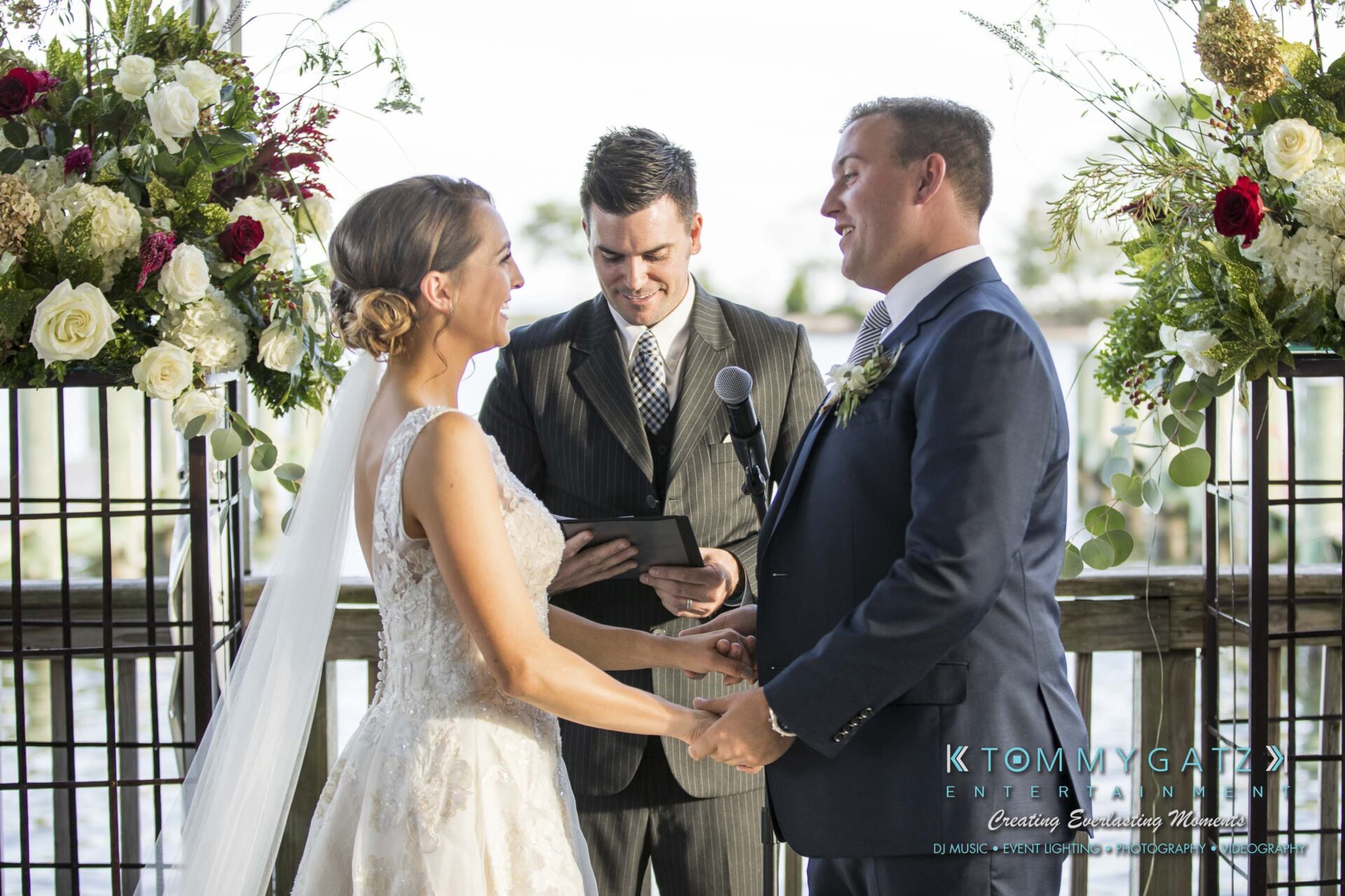 Amanda-Tyler-102117-CEREMONY-Low-Res-WM-67 - TOMMY GATZ ENTERTAINMENT A bride and groom hold hands during an outdoor wedding ceremony, with an officiant reading from a tablet. They're surrounded by flowers and decorative latticework.