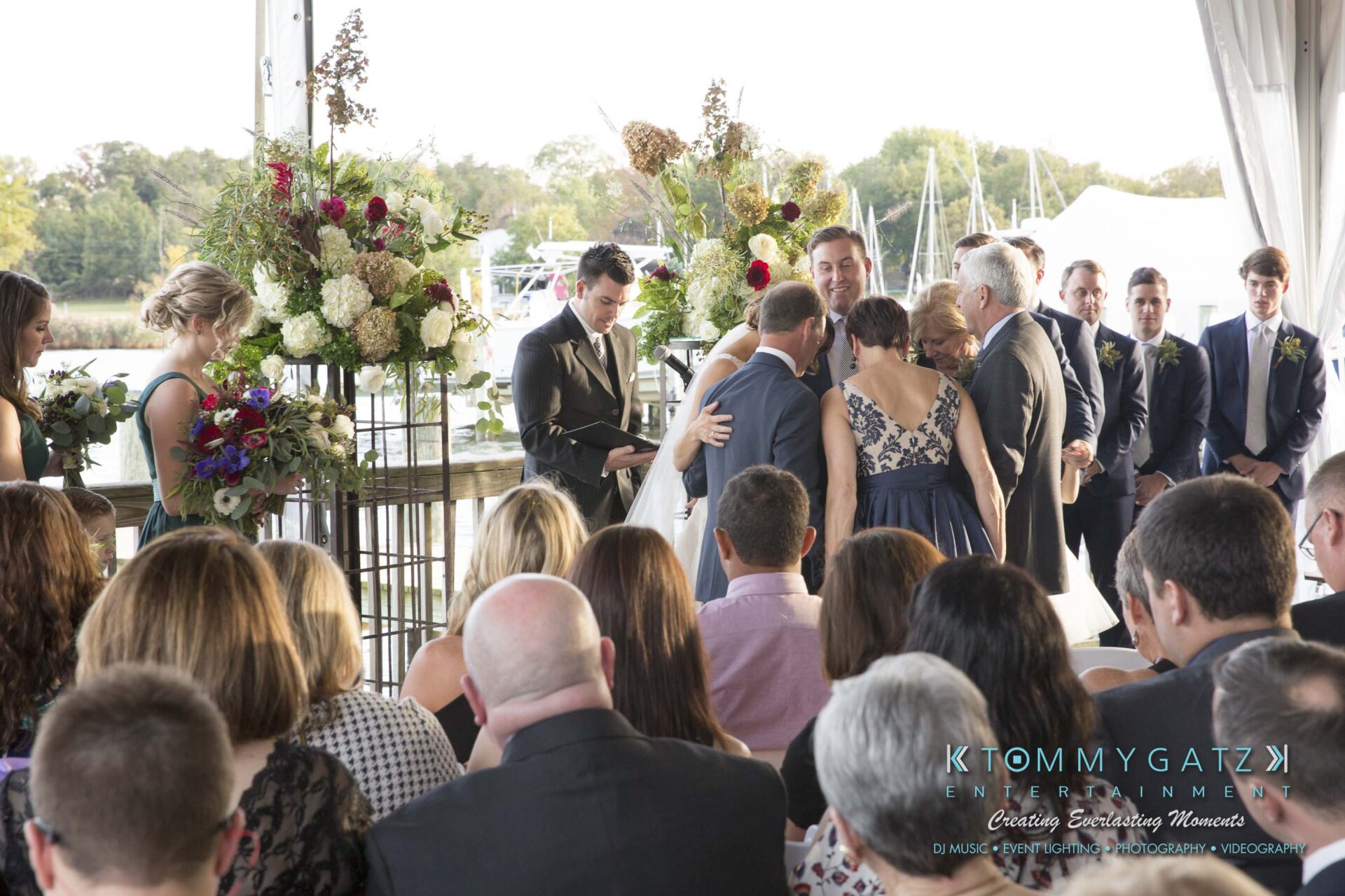 Amanda-Tyler-102117-CEREMONY-Low-Res-WM-76 - TOMMY GATZ ENTERTAINMENT Outdoor wedding ceremony by the water, with guests seated and a couple embracing near the altar surrounded by flowers and greenery.