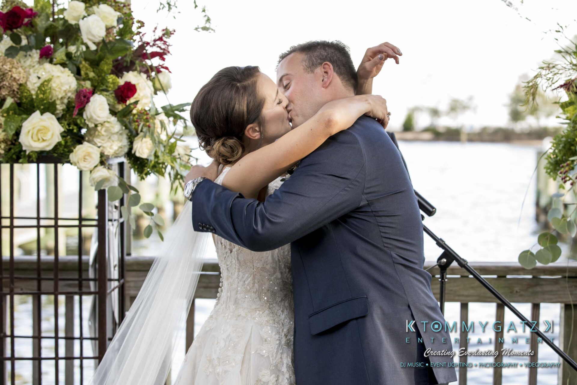Amanda-Tyler-102117-CEREMONY-Low-Res-WM-79 - TOMMY GATZ ENTERTAINMENT A bride and groom share a kiss at an outdoor wedding ceremony, surrounded by floral arrangements.