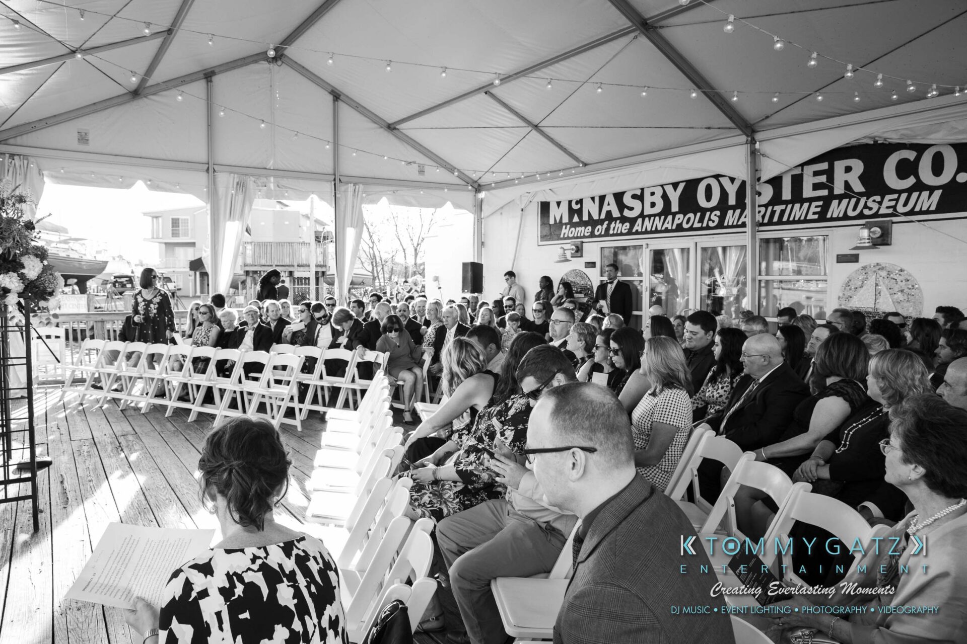 A black-and-white photo of people seated in rows under a tent with strings of lights above. A speaker stands at a podium. The backdrop has signage for M. Nasby Oyster Co. and the Annapolis Maritime Museum.
