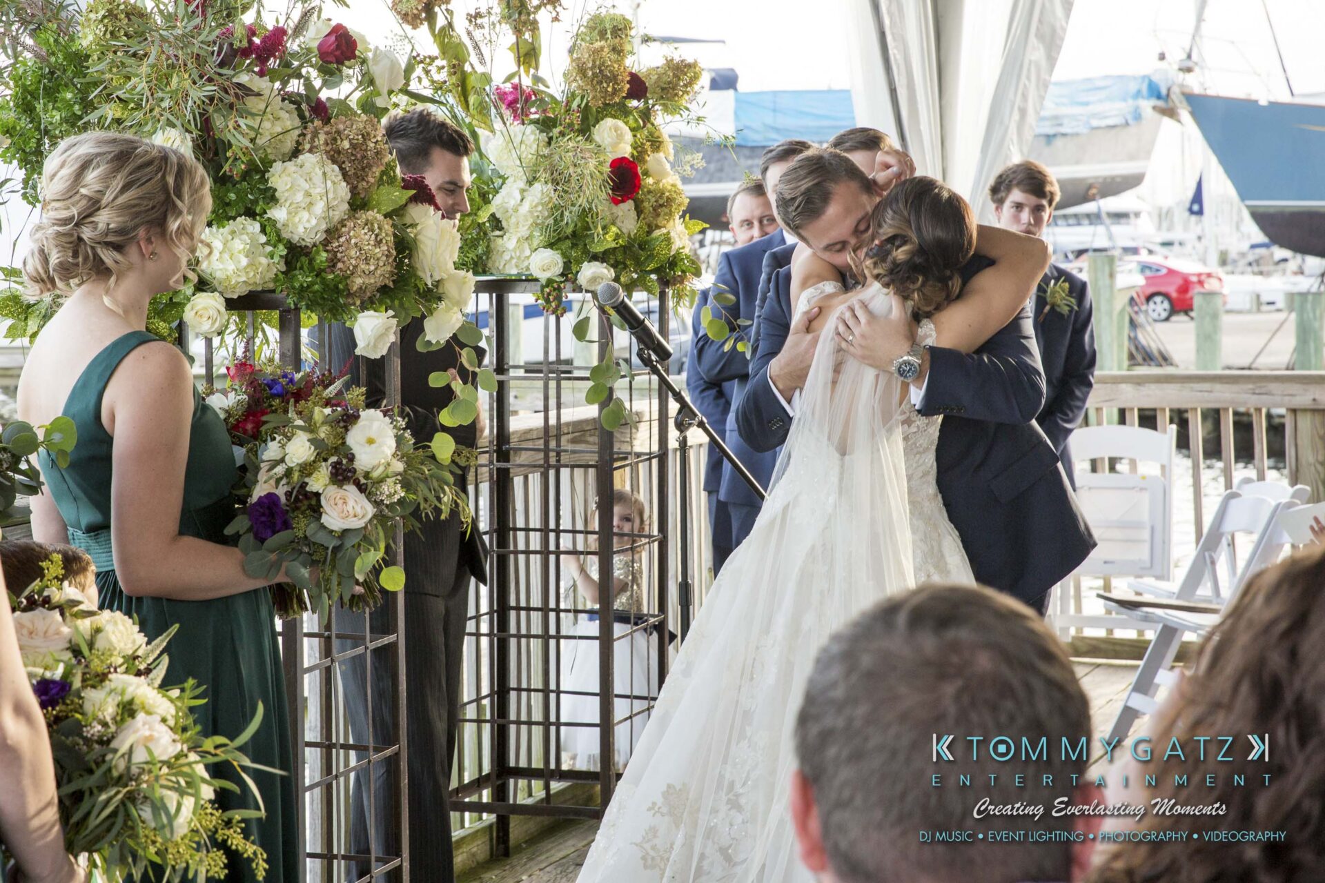 Amanda-Tyler-102117-CEREMONY-Low-Res-WM-80 - TOMMY GATZ ENTERTAINMENT A bride and groom embrace at an outdoor wedding ceremony, surrounded by bridesmaids and groomsmen, with floral arrangements and a boat in the background.