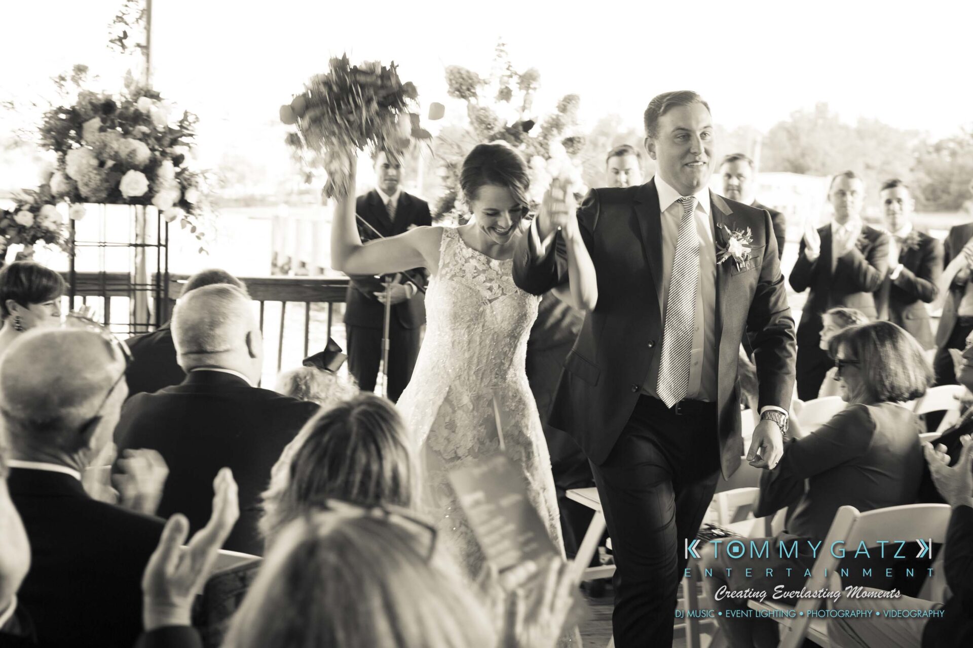 A bride and groom walk down the aisle, smiling and raising their hands, as guests applaud in a decorated venue.