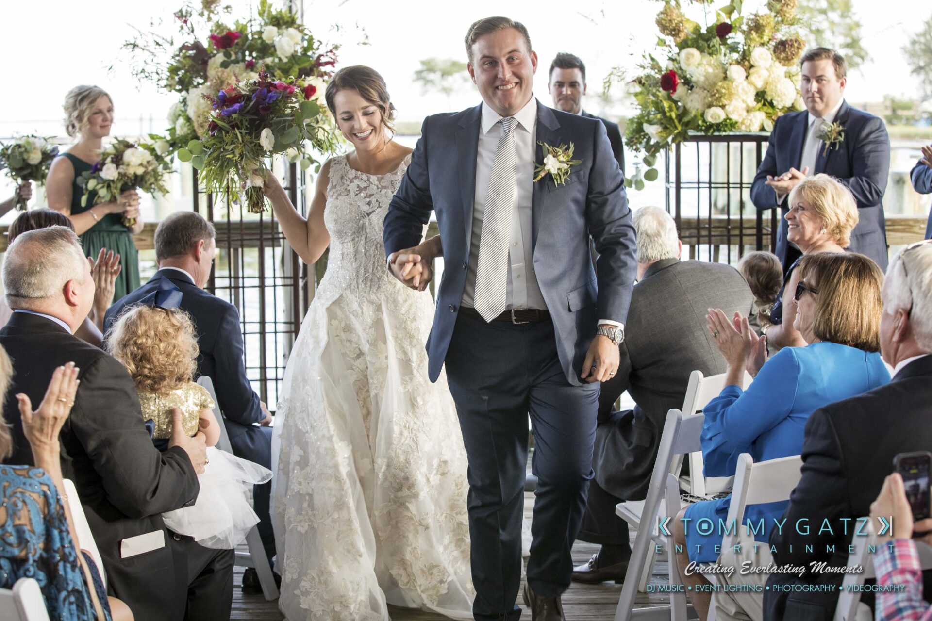 A bride and groom walk down the aisle, smiling, while guests clap. The setting is an outdoor area with floral decorations.