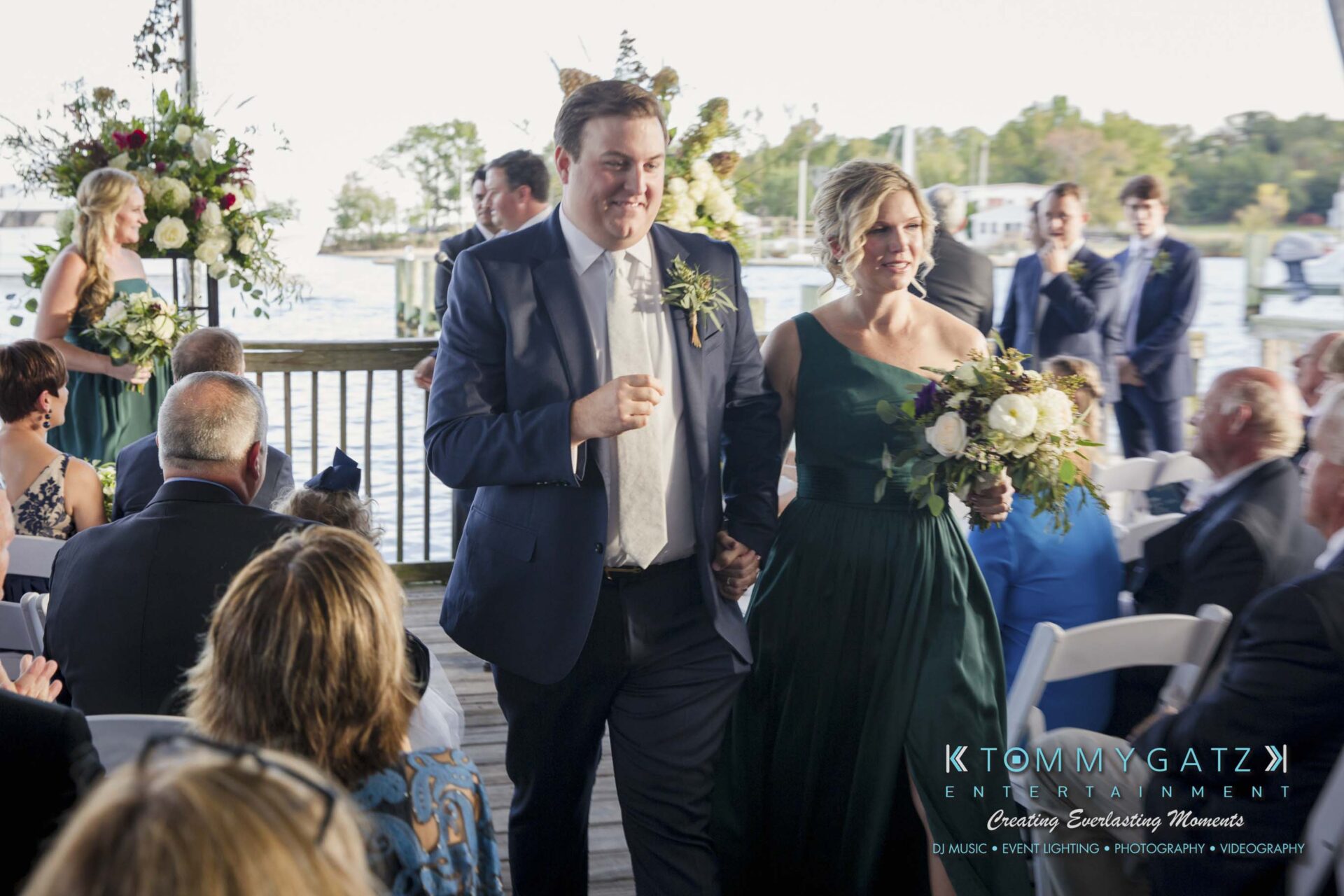 Amanda-Tyler-102117-CEREMONY-Low-Res-WM-90 - TOMMY GATZ ENTERTAINMENT A couple walks down an aisle at an outdoor event with guests seated. The woman carries a bouquet and both are dressed formally.