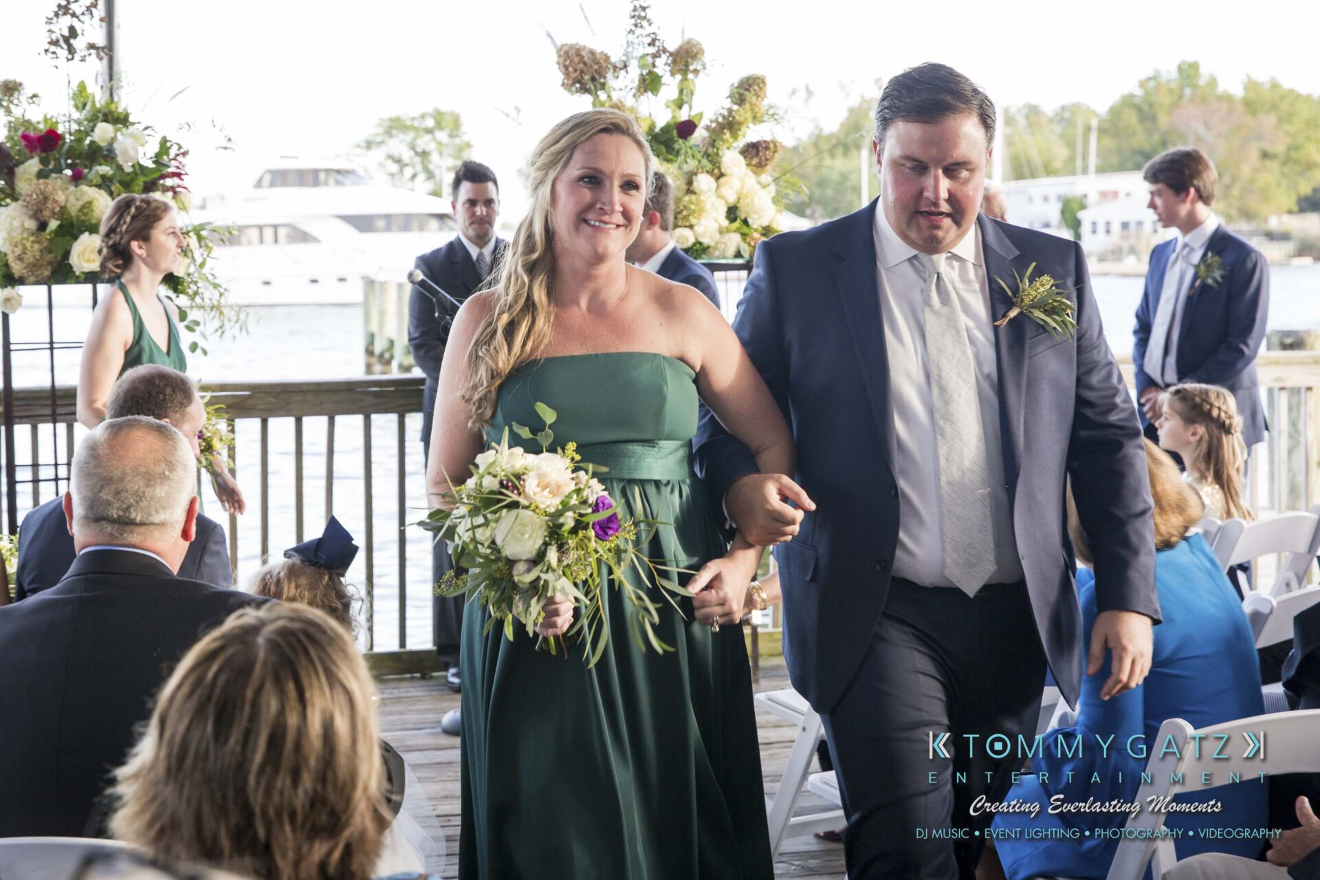 Amanda-Tyler-102117-CEREMONY-Low-Res-WM-92 - TOMMY GATZ ENTERTAINMENT A couple walks down the aisle at an outdoor wedding ceremony, with the bride holding a bouquet and wearing a green dress, and the groom in a suit, surrounded by guests and floral decorations.