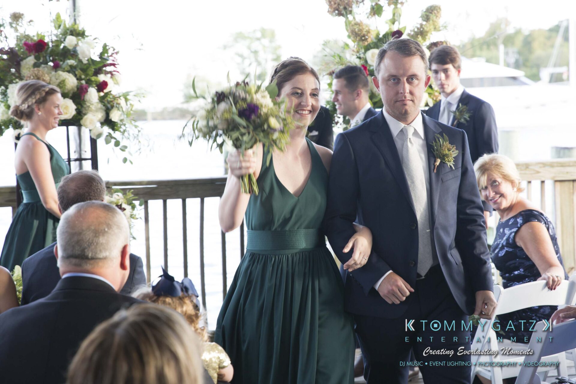 Amanda-Tyler-102117-CEREMONY-Low-Res-WM-93 - TOMMY GATZ ENTERTAINMENT A couple walks down the aisle at an outdoor wedding ceremony. The woman holds a bouquet and smiles, while the man has a neutral expression. Guests are seated on either side.