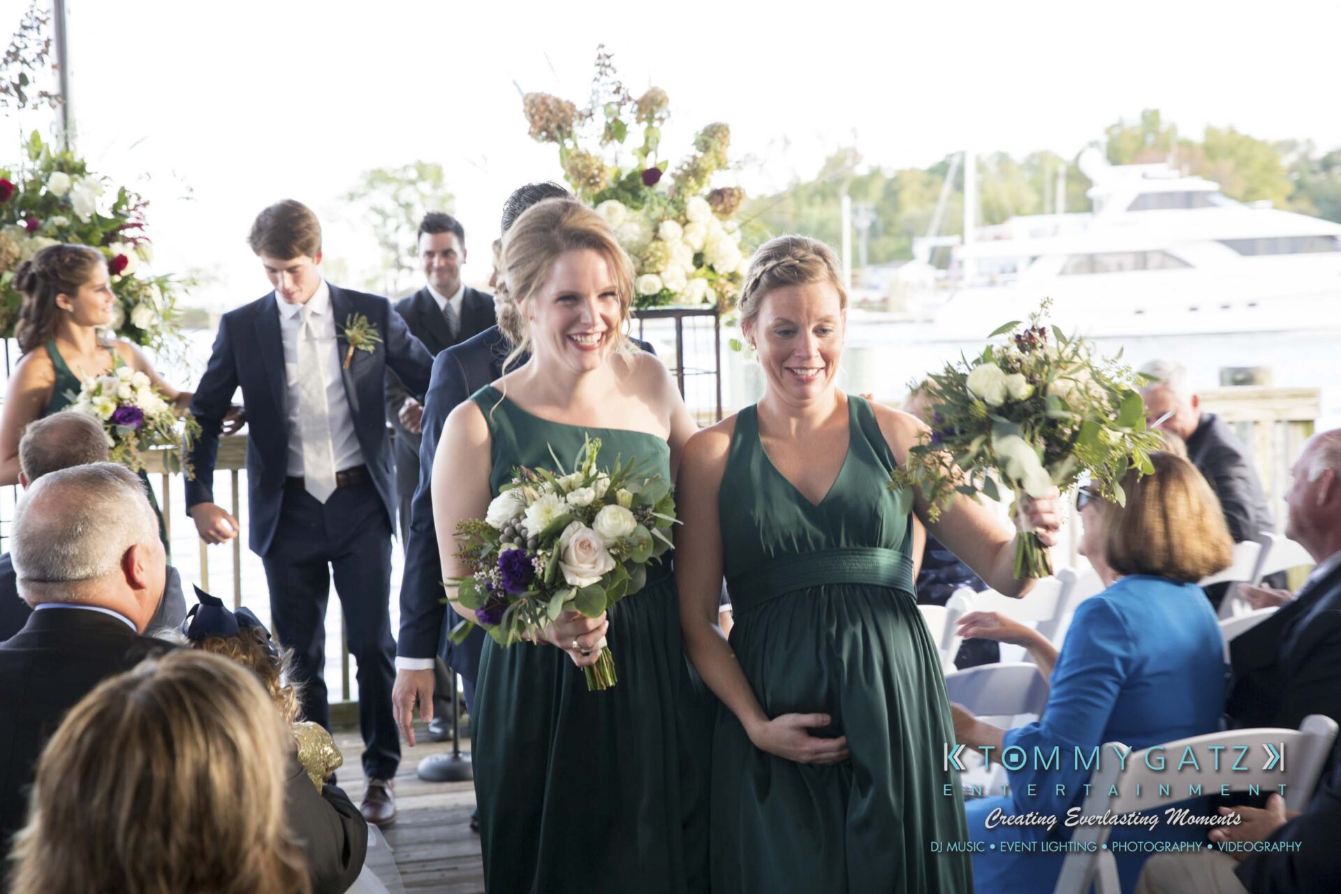 Two women in green dresses hold bouquets at an outdoor event by the water. A group of people is gathered, and a white boat is in the background.