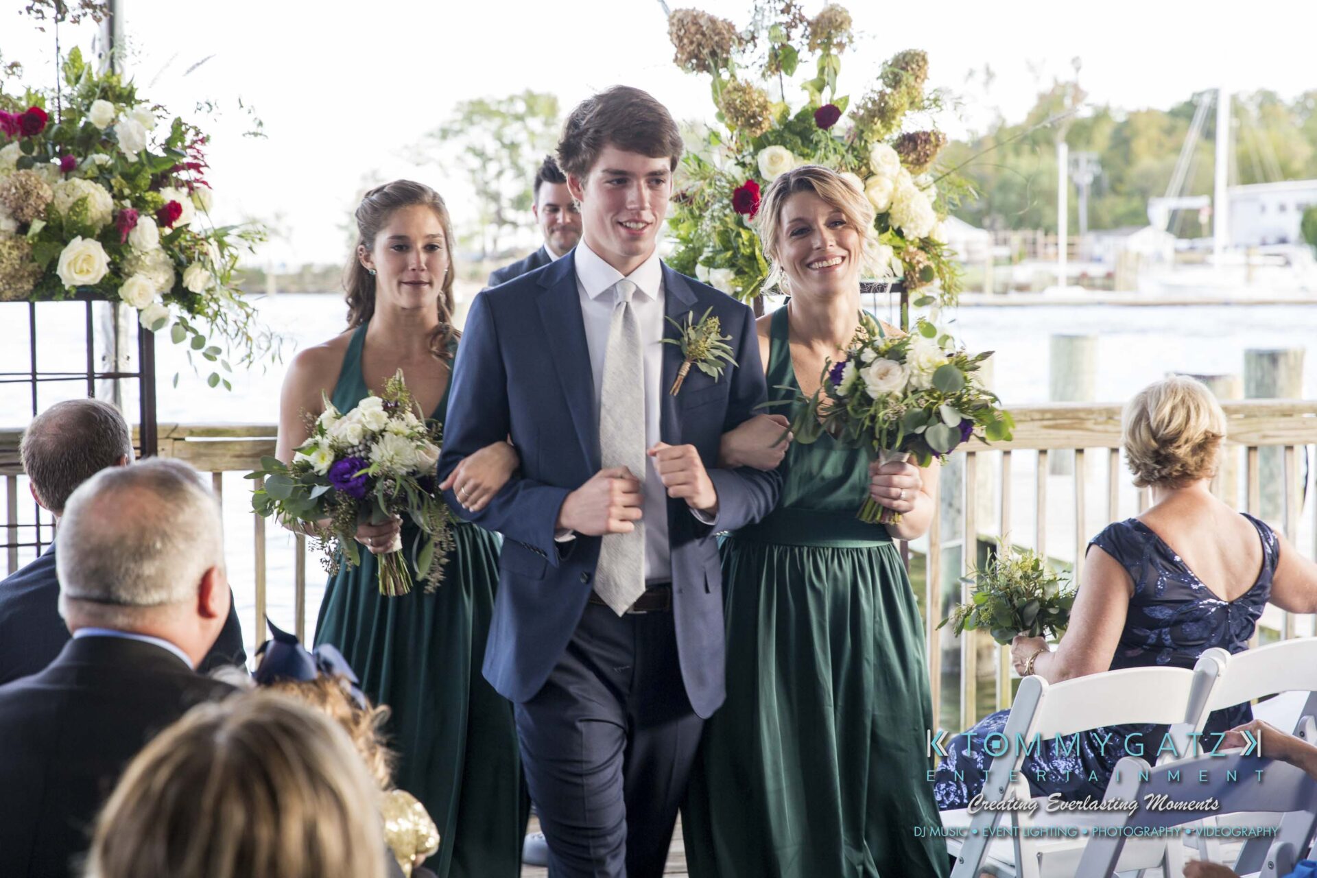 A young man in a suit walks down an aisle with two women in green dresses, carrying bouquets, at an outdoor wedding ceremony by the water. Guests are seated on either side.