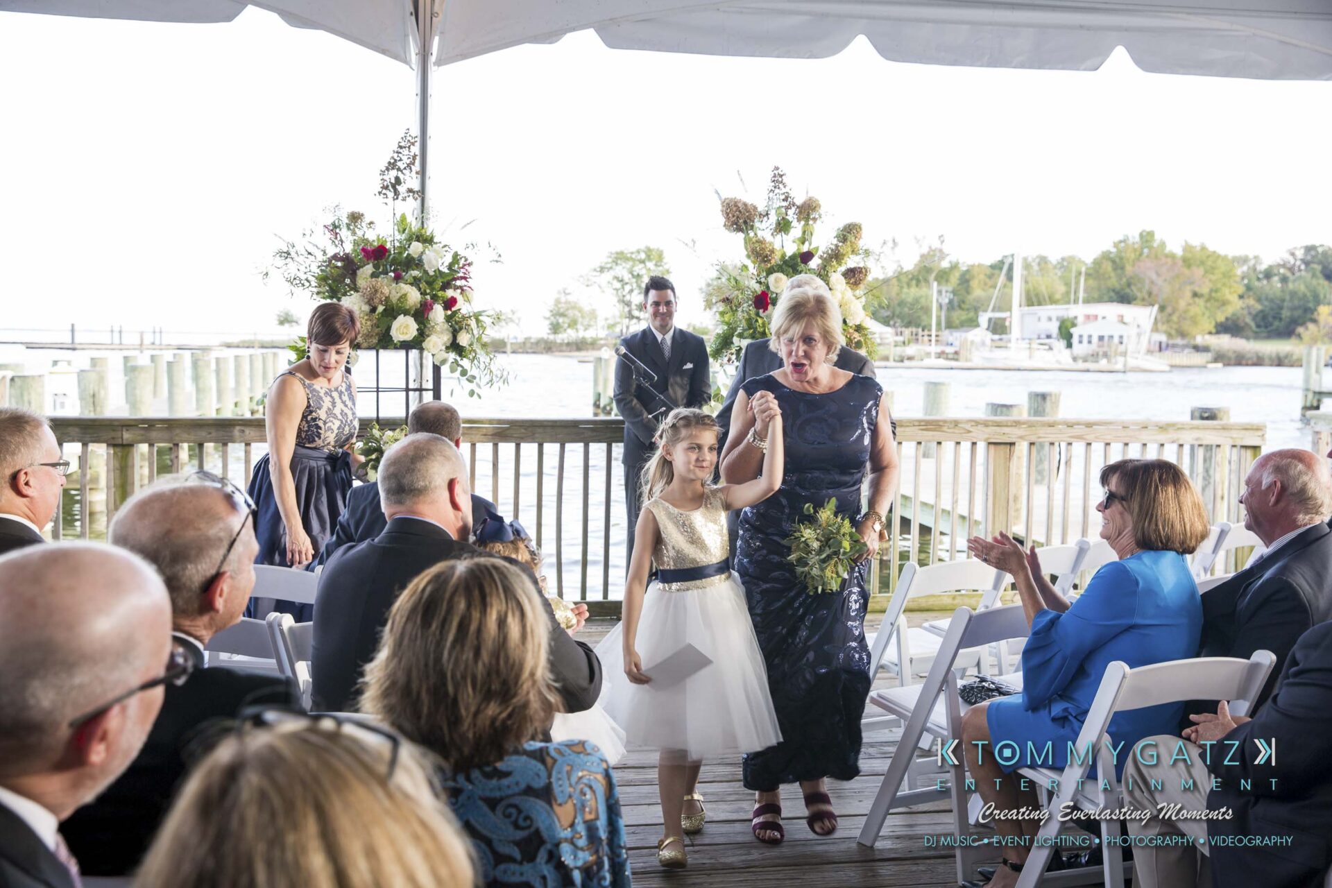 Young girl and older woman walk down the aisle under a canopy at an outdoor wedding ceremony. Guests are seated on either side.