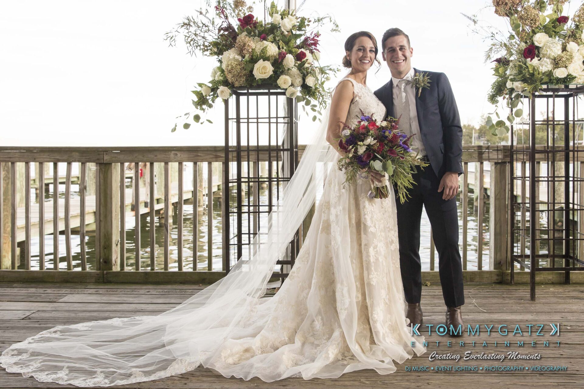 Bride and groom smiling at the camera on a wooden deck with floral decorations. Bride holds a bouquet and wears a gown with a long veil.