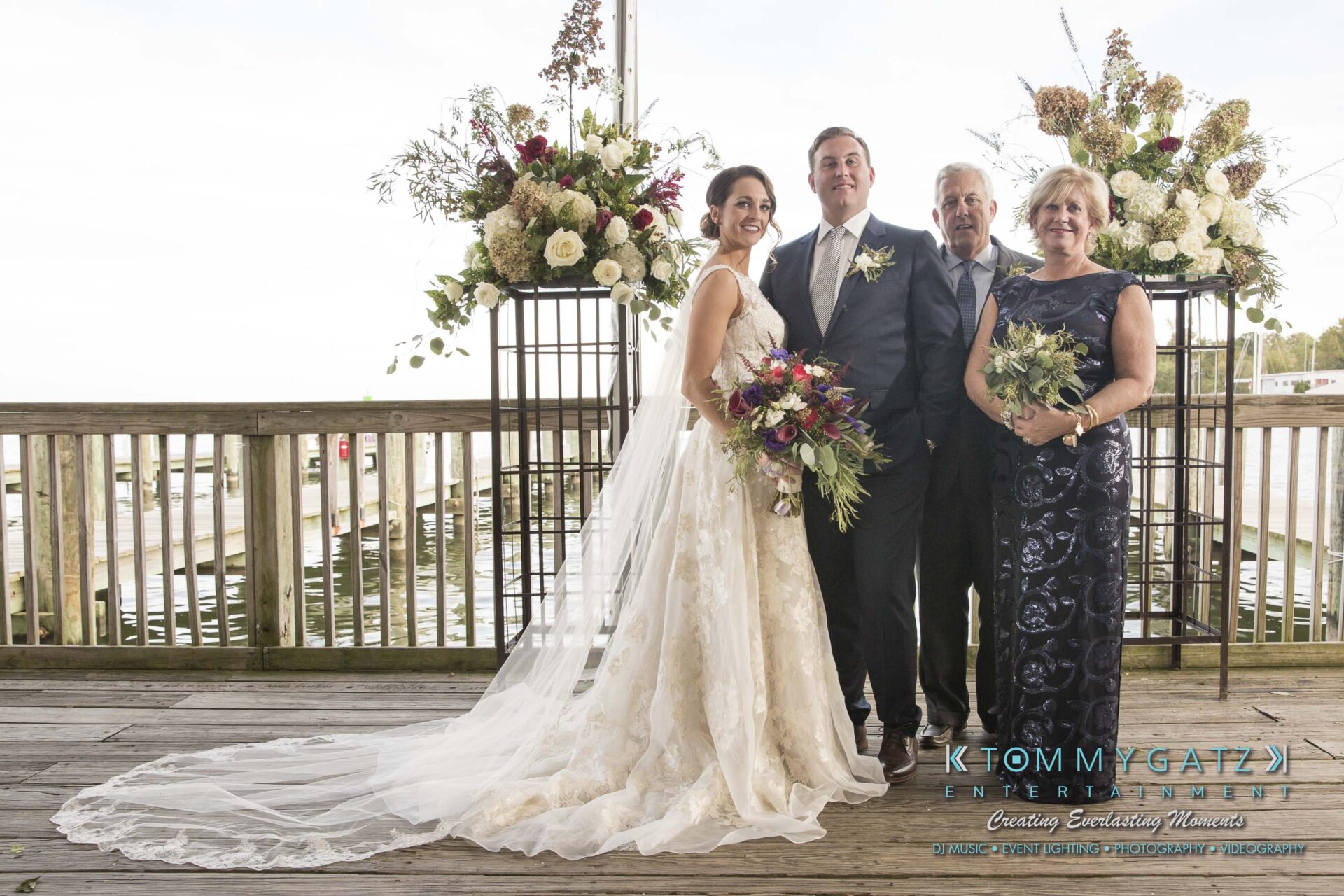 A bride and groom stand on a wooden deck with two older adults, likely parents. They are all smiling, holding bouquets, with floral arrangements and a waterfront in the background.