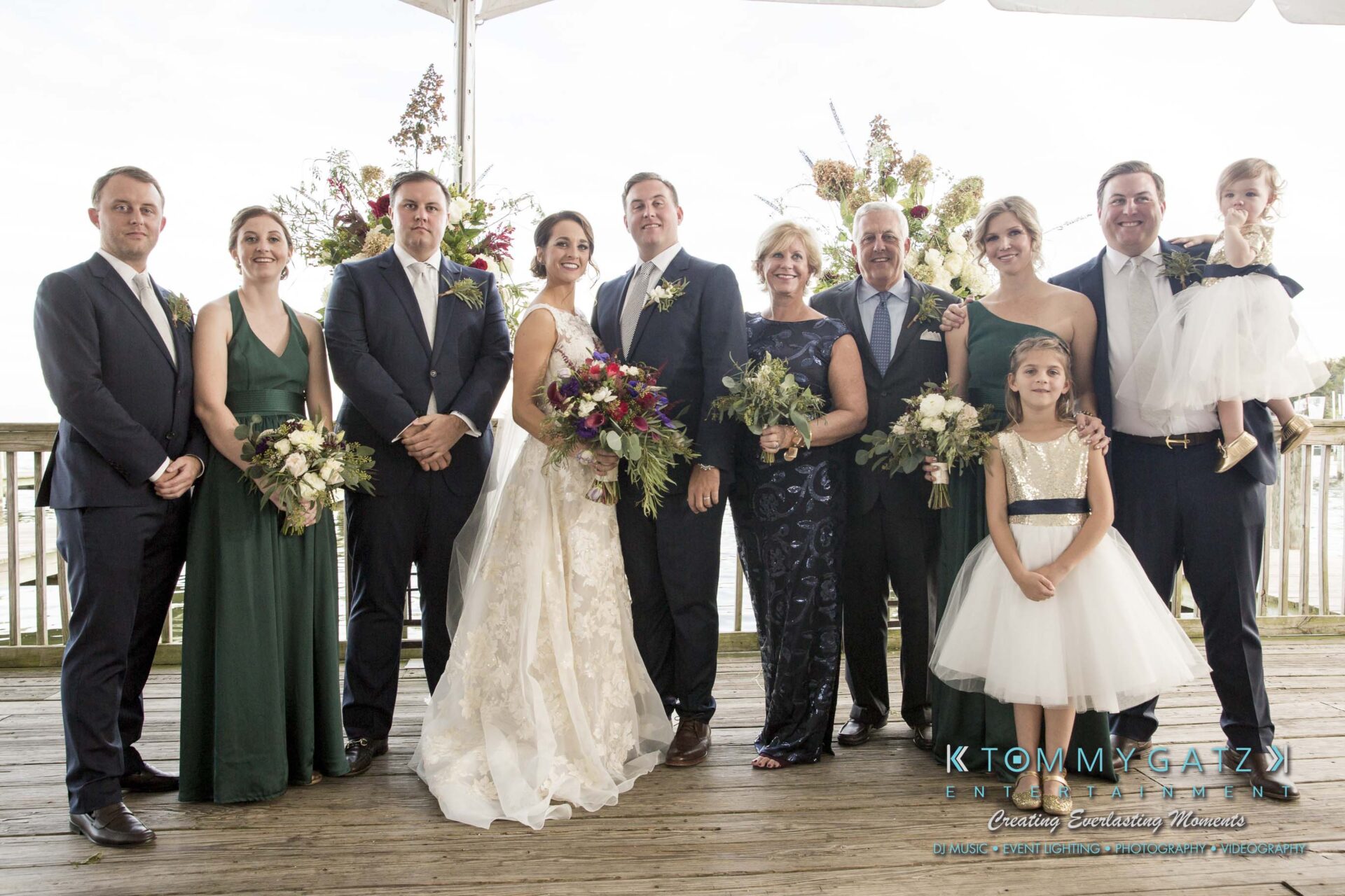 A wedding party poses on a wooden deck. The bride and groom stand in the center, surrounded by family and friends, all dressed in formal attire. Two flower girls are in front.