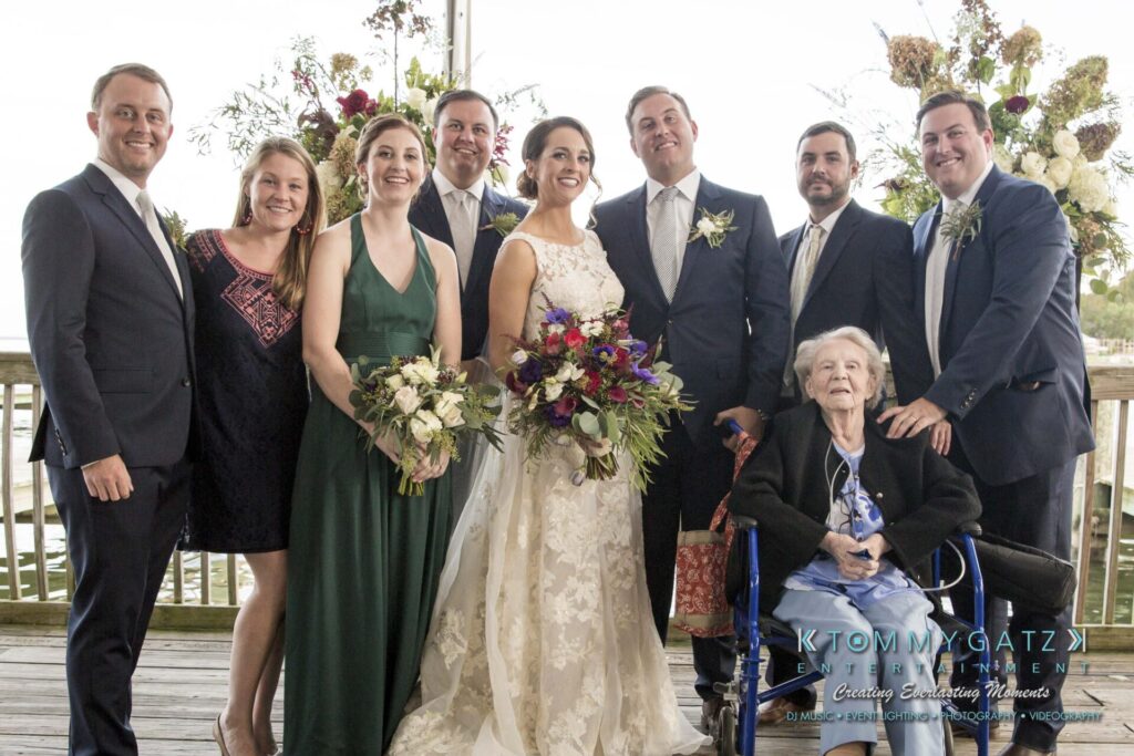 Amanda-Tyler-102117-FORMALS-Low-Res-WM-15 - TOMMY GATZ ENTERTAINMENT A group photo with eight people standing and one person seated in a wheelchair, all dressed formally, on a wooden deck with floral decorations in the background.