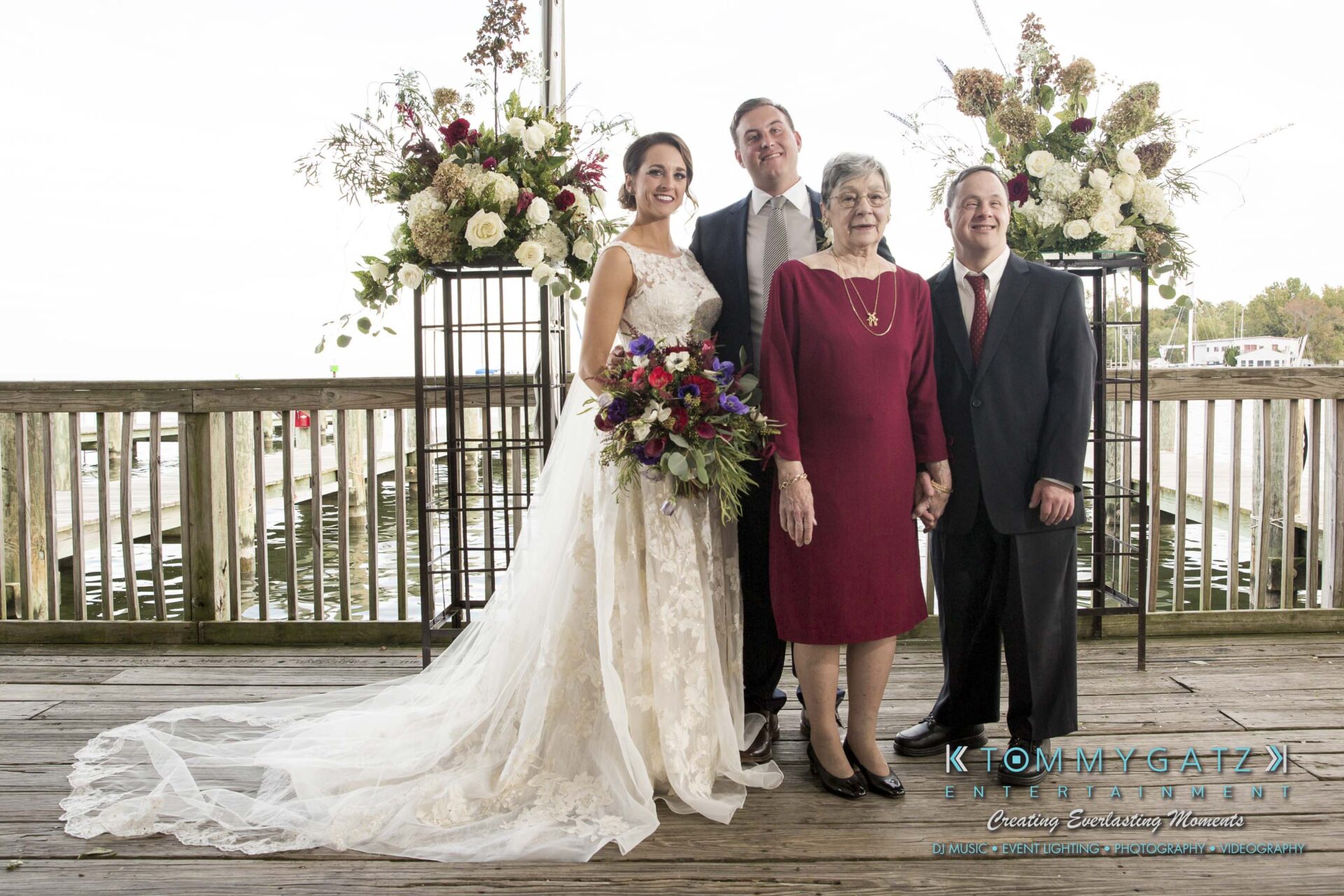 Amanda-Tyler-102117-FORMALS-Low-Res-WM-17 - TOMMY GATZ ENTERTAINMENT A bride and groom pose with two older adults on a wooden deck, with floral arrangements in the background.