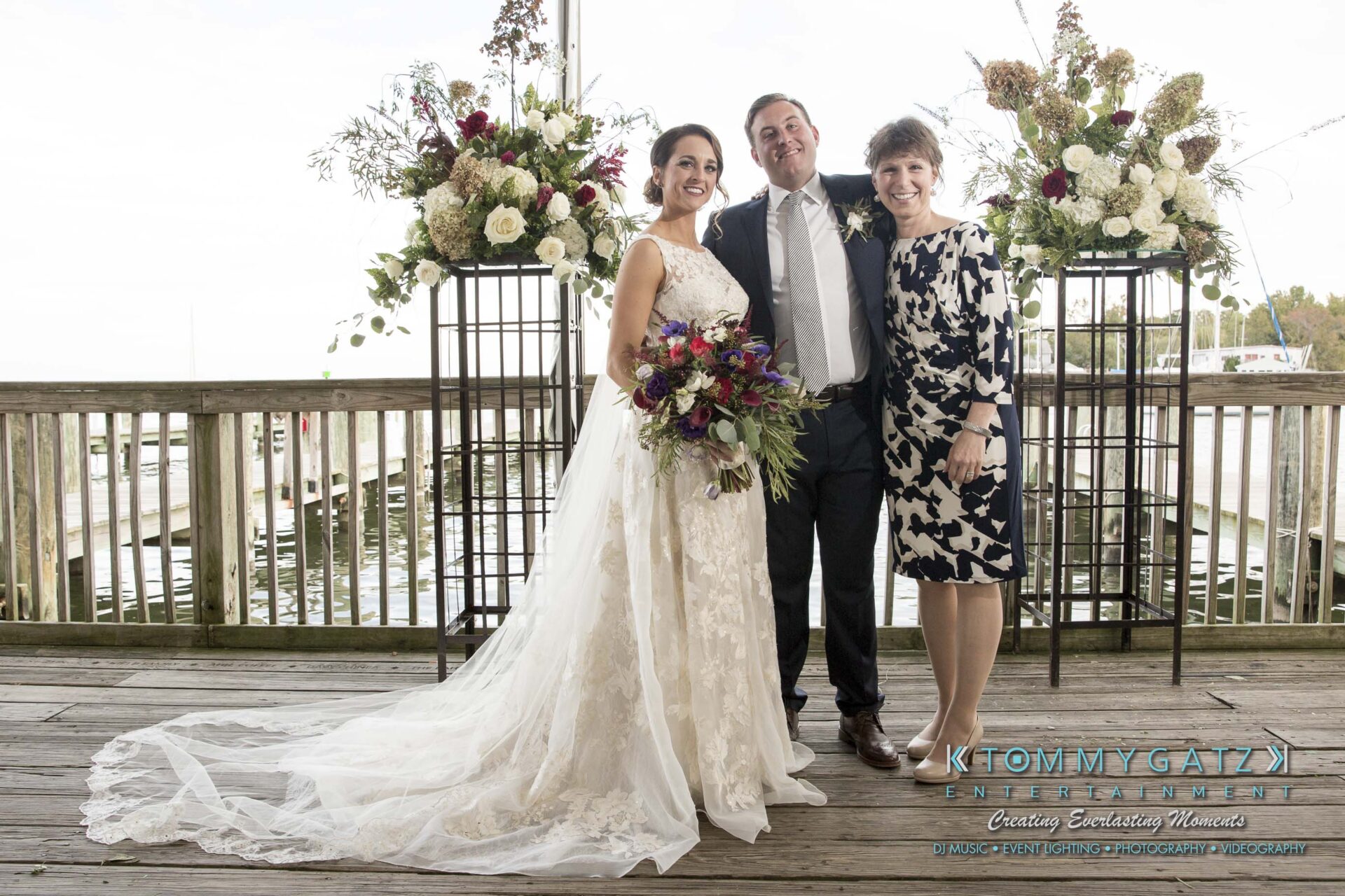 Amanda-Tyler-102117-FORMALS-Low-Res-WM-18 - TOMMY GATZ ENTERTAINMENT A bride and groom pose with a woman on a deck decorated with floral arrangements.