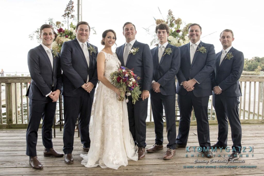 Amanda-Tyler-102117-FORMALS-Low-Res-WM-19 - TOMMY GATZ ENTERTAINMENT A bride and groom pose with five groomsmen on a wooden deck, with floral arrangements and water in the background.