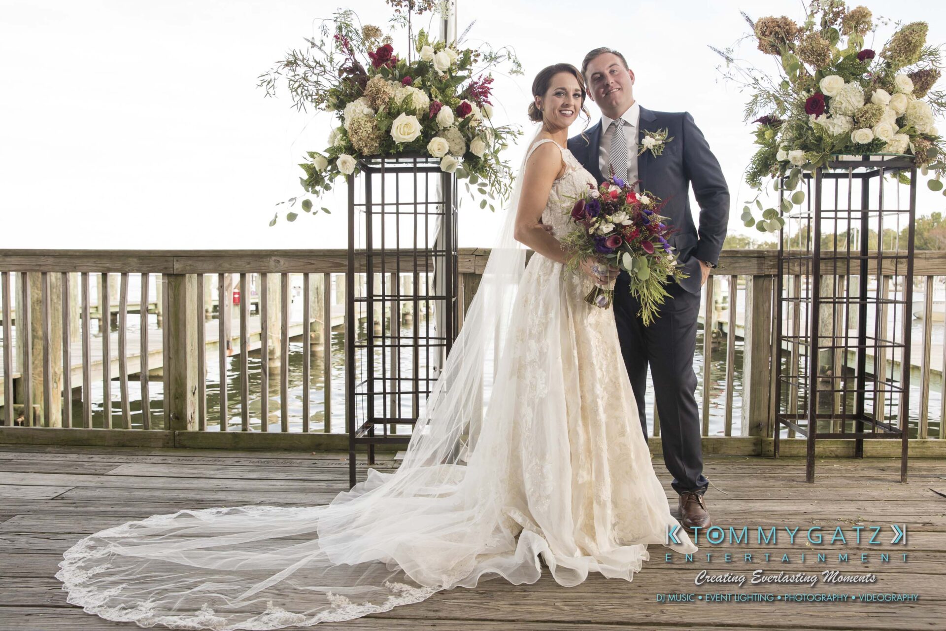 Amanda-Tyler-102117-FORMALS-Low-Res-WM-2 - TOMMY GATZ ENTERTAINMENT A bride and groom pose together on a wooden deck, surrounded by floral arrangements. The bride wears a lace gown with a long train, and the groom is in a suit and tie.