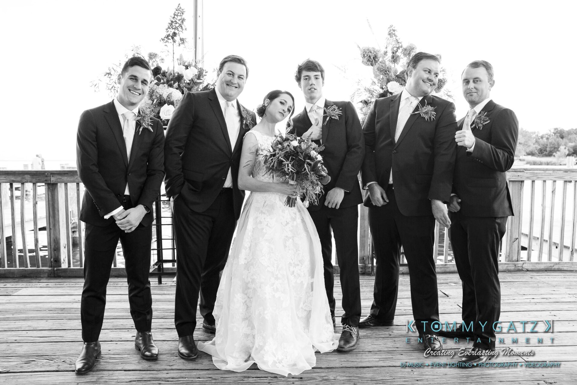 Amanda-Tyler-102117-FORMALS-Low-Res-WM-21 - TOMMY GATZ ENTERTAINMENT Bride and groom pose with groomsmen on a wooden deck, some smiling and gesturing, with floral arrangements in the background.