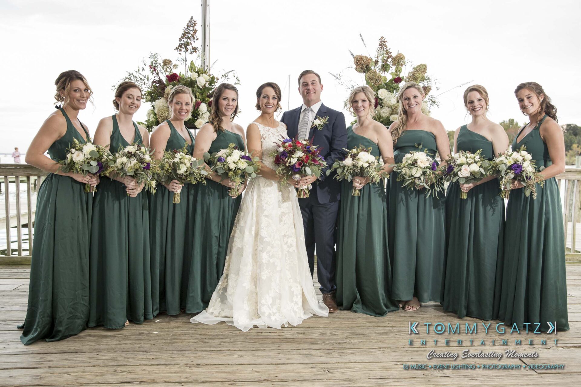 Amanda-Tyler-102117-FORMALS-Low-Res-WM-22 - TOMMY GATZ ENTERTAINMENT A bride and groom pose with eight bridesmaids in green dresses on a wooden deck, each holding a bouquet.