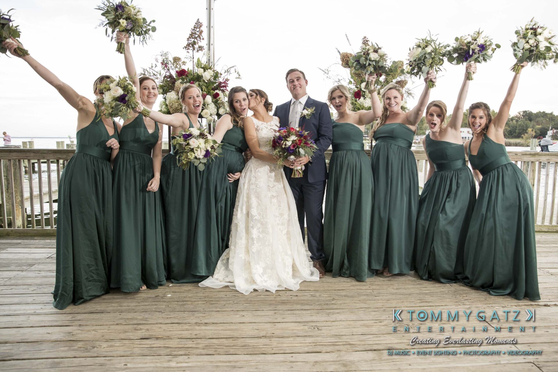 Amanda-Tyler-102117-FORMALS-Low-Res-WM-24 - TOMMY GATZ ENTERTAINMENT A bride and groom stand on a wooden deck surrounded by eight bridesmaids in green dresses, all holding bouquets and raising their arms in celebration.