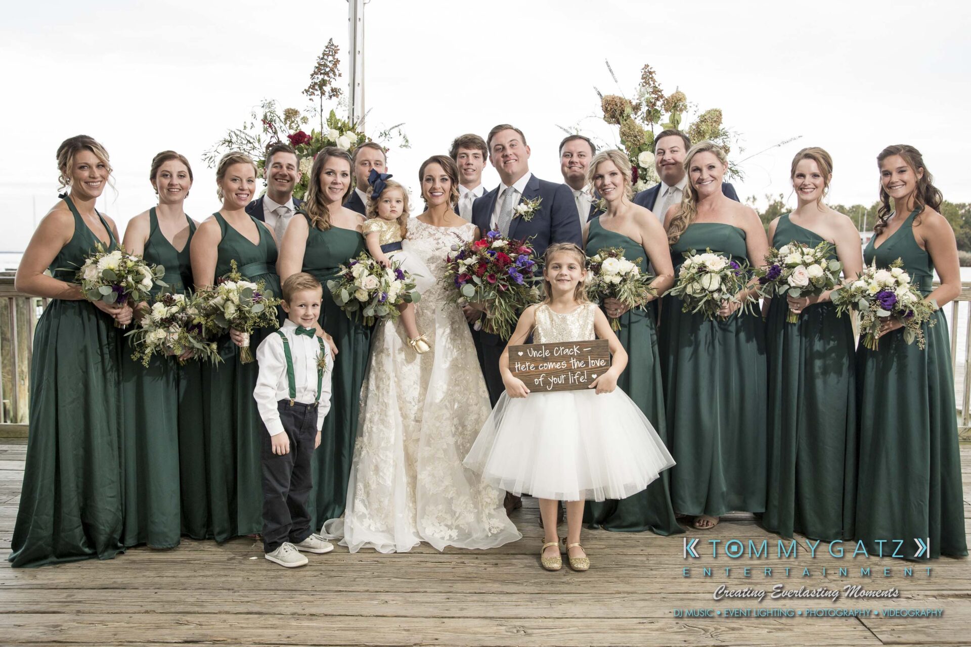 Amanda-Tyler-102117-FORMALS-Low-Res-WM-26 - TOMMY GATZ ENTERTAINMENT A wedding party poses on a wooden deck. The bride and groom stand in the center, surrounded by bridesmaids in green dresses and groomsmen in suits. Two children are at the front holding signs.