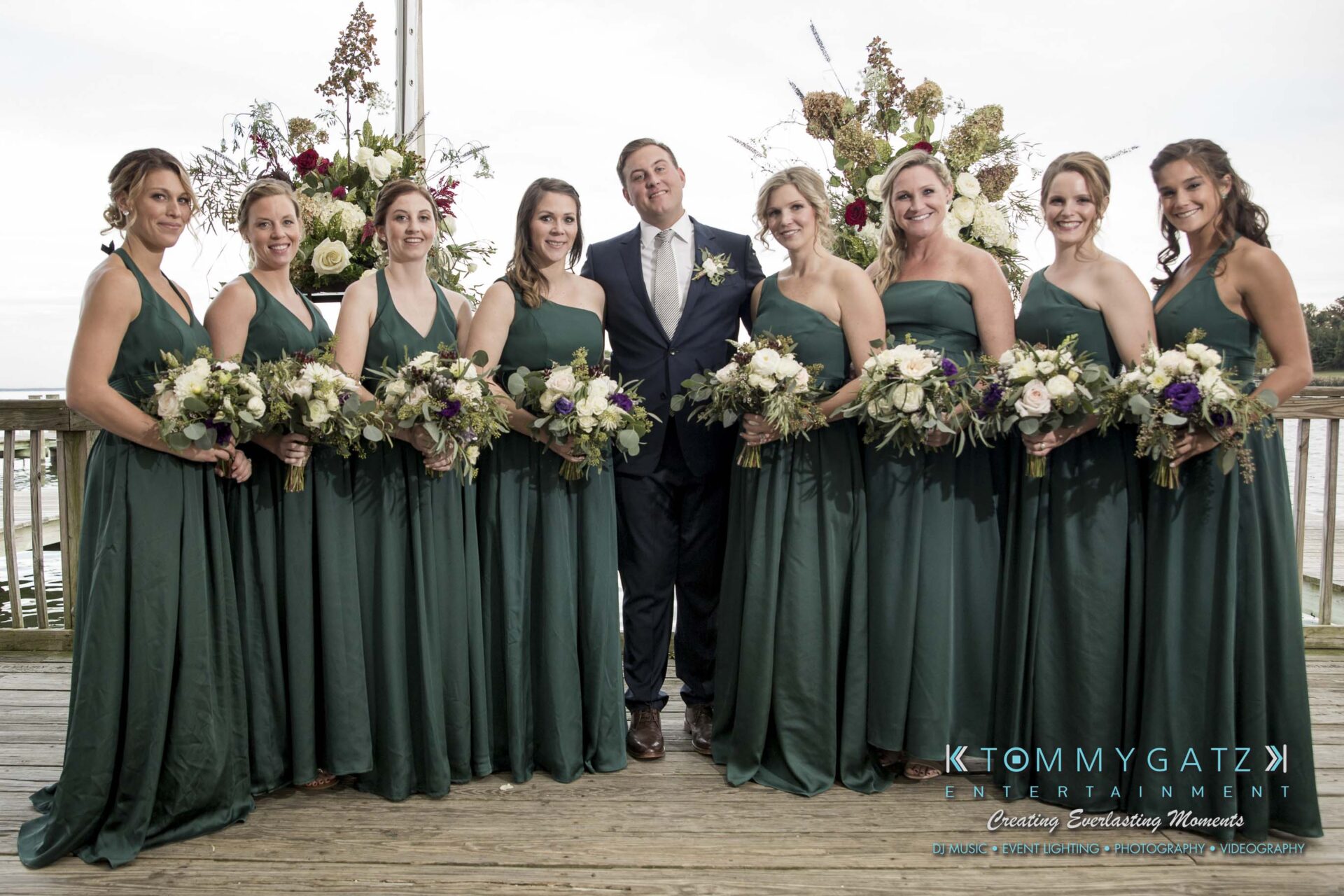 Amanda-Tyler-102117-FORMALS-Low-Res-WM-28 - TOMMY GATZ ENTERTAINMENT A groom stands with eight bridesmaids in green dresses, all holding bouquets, on a wooden deck.