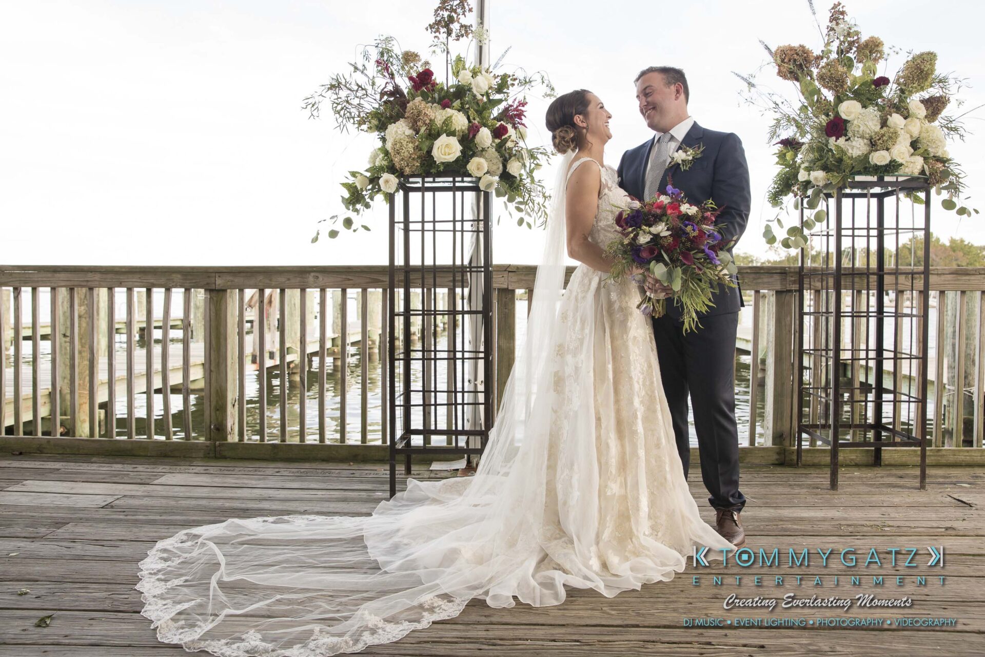 Amanda-Tyler-102117-FORMALS-Low-Res-WM-3 - TOMMY GATZ ENTERTAINMENT A bride and groom stand together on a wooden deck, surrounded by floral arrangements, with water and greenery in the background.