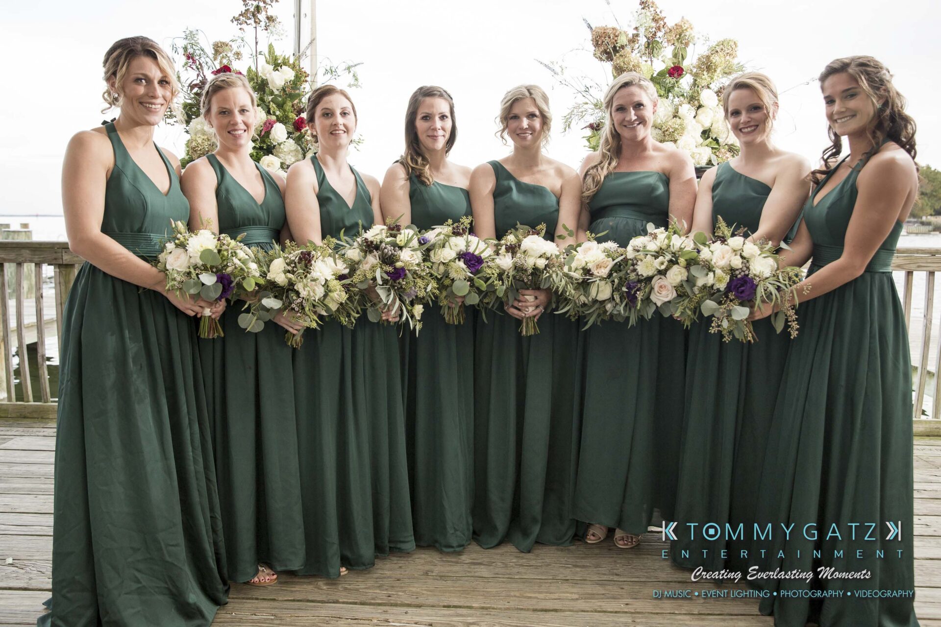 Amanda-Tyler-102117-FORMALS-Low-Res-WM-30 - TOMMY GATZ ENTERTAINMENT Eight bridesmaids in dark green dresses stand in a row holding floral bouquets on a wooden deck.