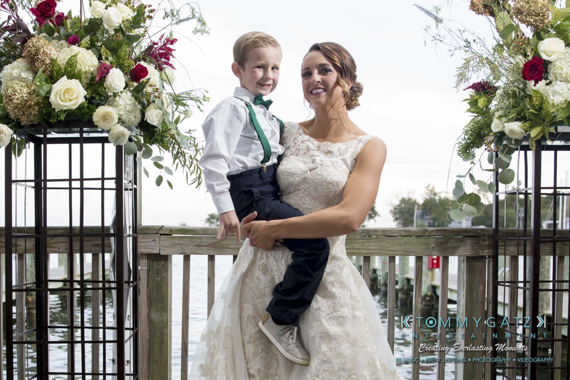 Amanda-Tyler-102117-FORMALS-Low-Res-WM-31 - TOMMY GATZ ENTERTAINMENT A woman in a white dress holds a young boy wearing a white shirt and bow tie, standing on a deck with flowers and a body of water in the background.