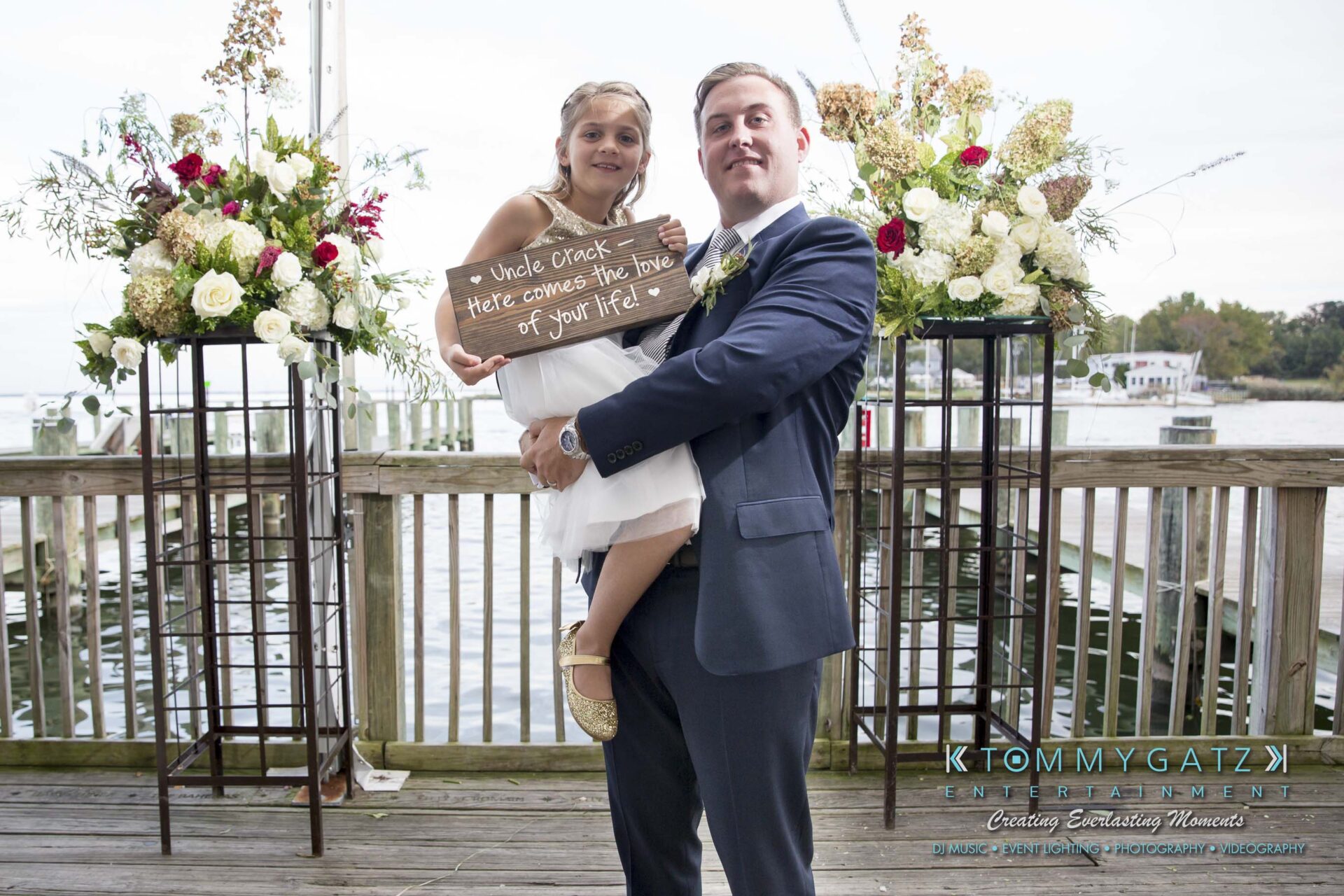Amanda-Tyler-102117-FORMALS-Low-Res-WM-33 - TOMMY GATZ ENTERTAINMENT A man in a suit holds a child in a white dress. The child holds a sign reading, "Uncle (name), here comes the love of your life!" Flowers and water are in the background.