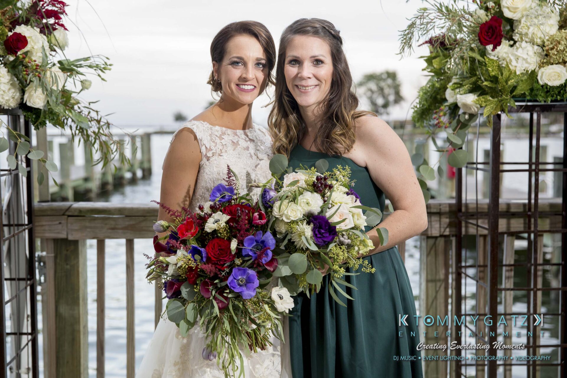 Amanda-Tyler-102117-FORMALS-Low-Res-WM-38 - TOMMY GATZ ENTERTAINMENT Two women in formal dresses smile while holding colorful flower bouquets. They stand on a wooden deck with water in the background.