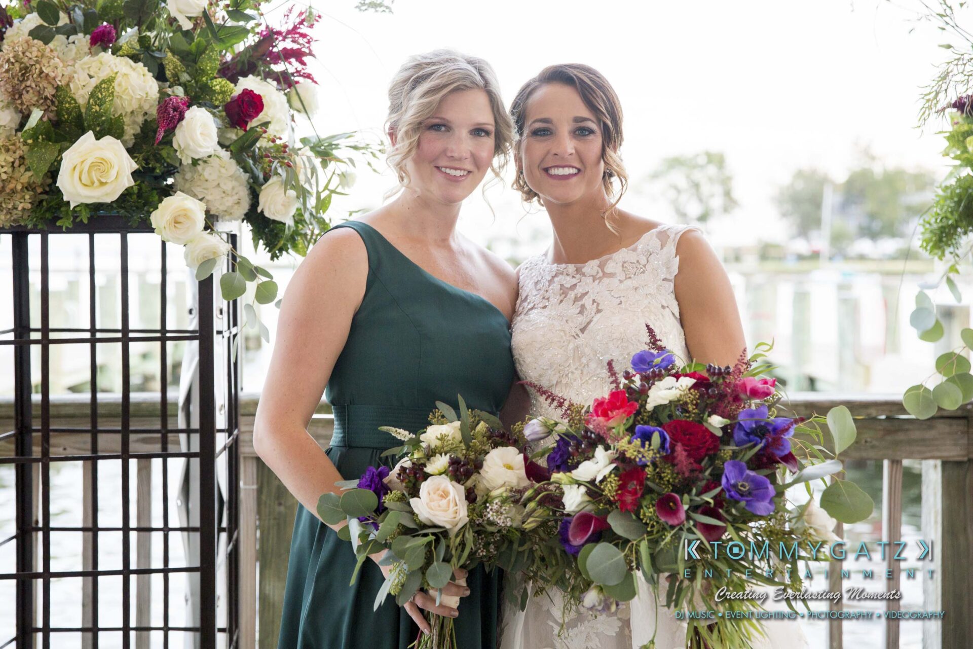 Amanda-Tyler-102117-FORMALS-Low-Res-WM-39 - TOMMY GATZ ENTERTAINMENT Two women, one in a white dress and the other in a green dress, smiling and holding flower bouquets in front of a wooden railing with greenery and flowers.