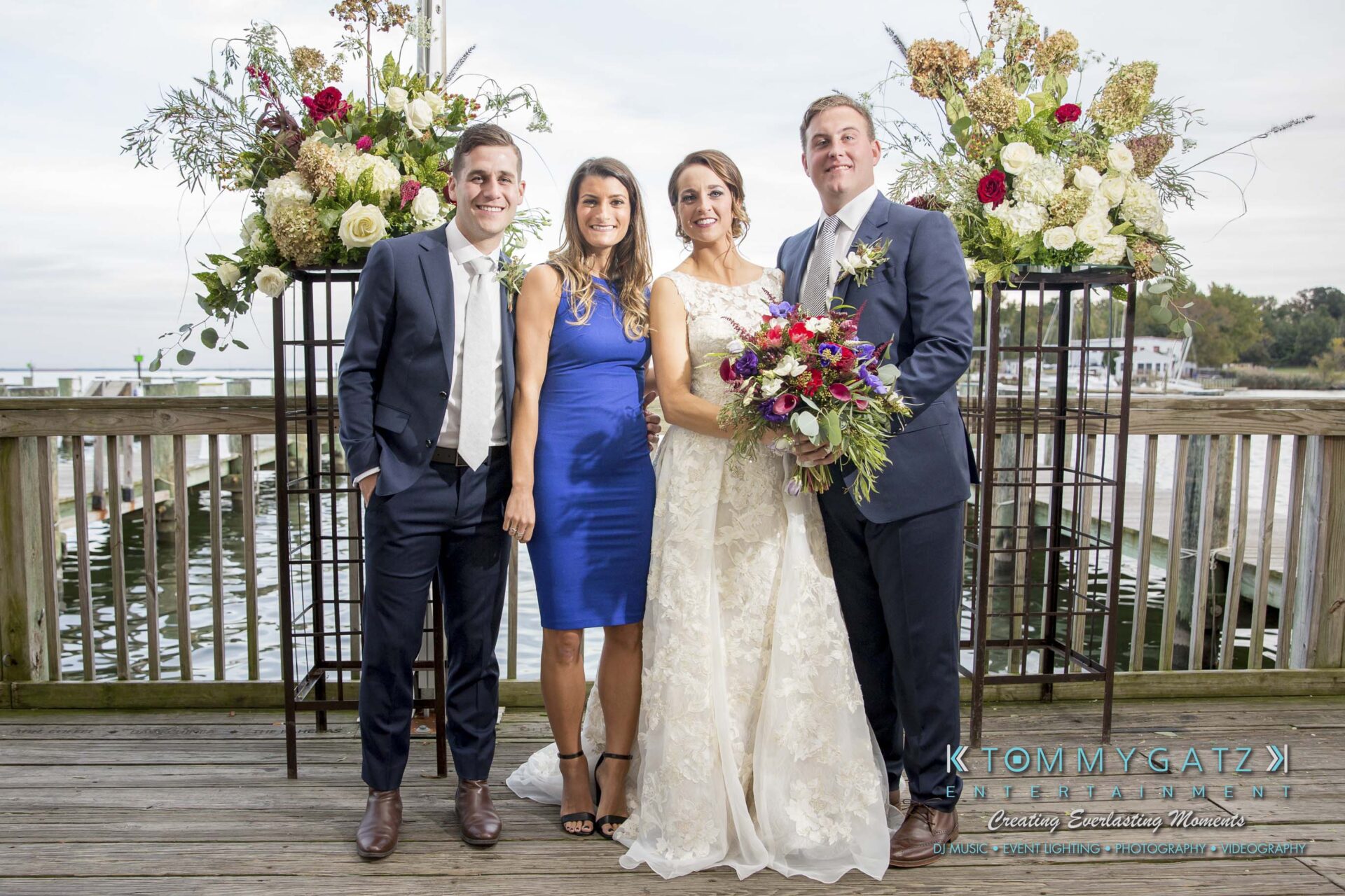 Amanda-Tyler-102117-FORMALS-Low-Res-WM-40 - TOMMY GATZ ENTERTAINMENT Four people stand on a wooden deck, including a bride and groom in wedding attire, flanked by two guests. Ornate flower arrangements are in the background.