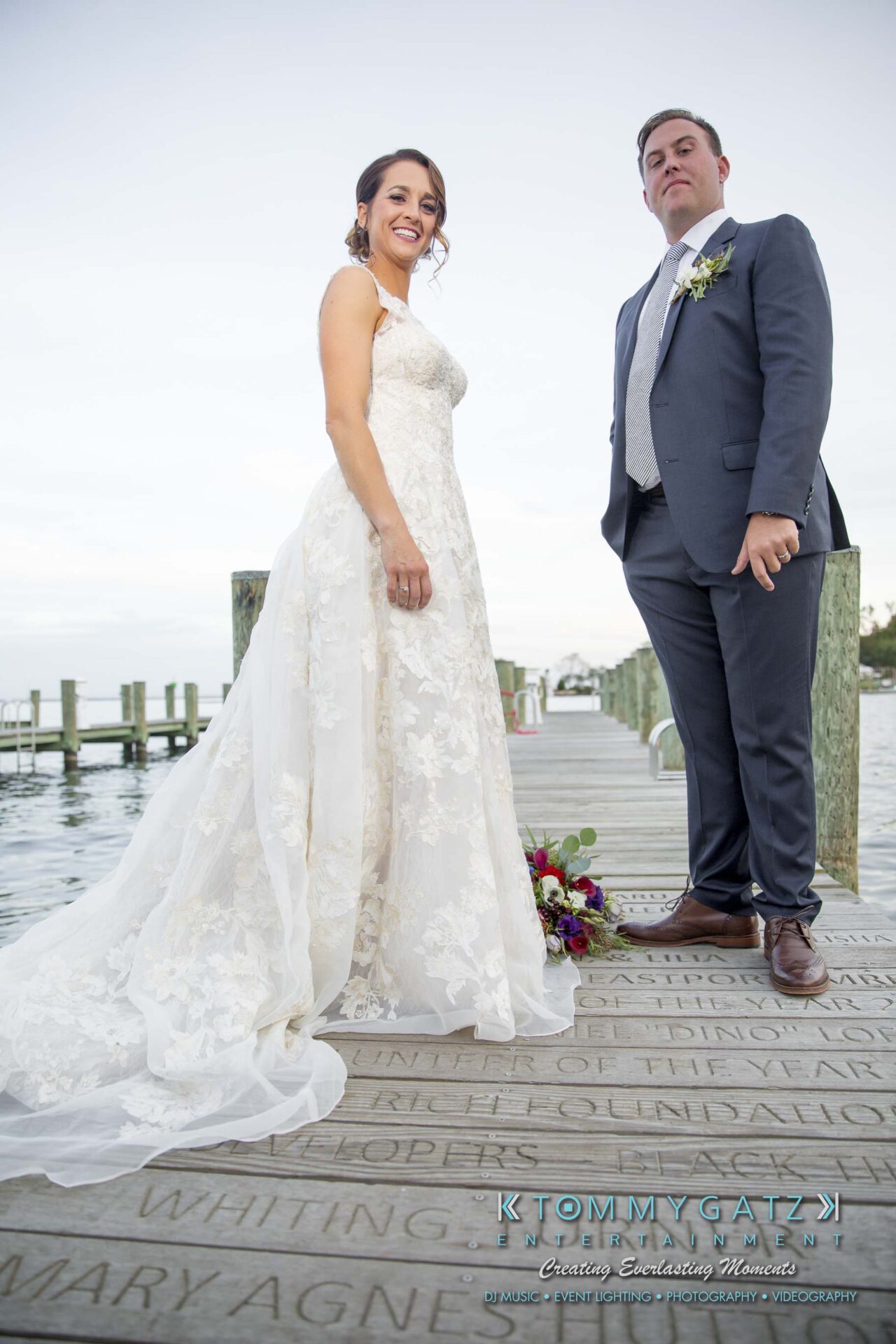 Amanda-Tyler-102117-FORMALS-Low-Res-WM-44 - TOMMY GATZ ENTERTAINMENT A bride and groom stand on a wooden dock by the water. The bride wears a lace gown, and the groom is in a suit. A bouquet is on the dock.