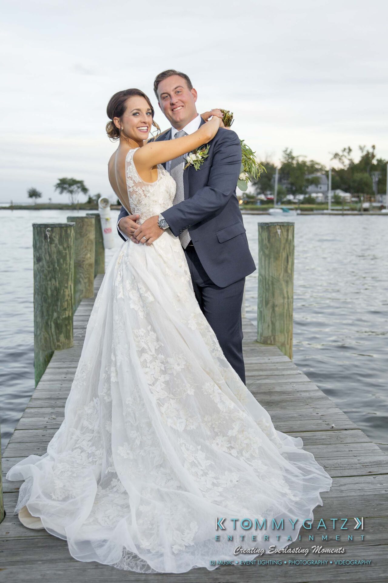 Amanda-Tyler-102117-FORMALS-Low-Res-WM-45 - TOMMY GATZ ENTERTAINMENT Bride and groom stand on a dock by the water, smiling as they embrace. The bride wears a lace dress, and the groom is in a blue suit.