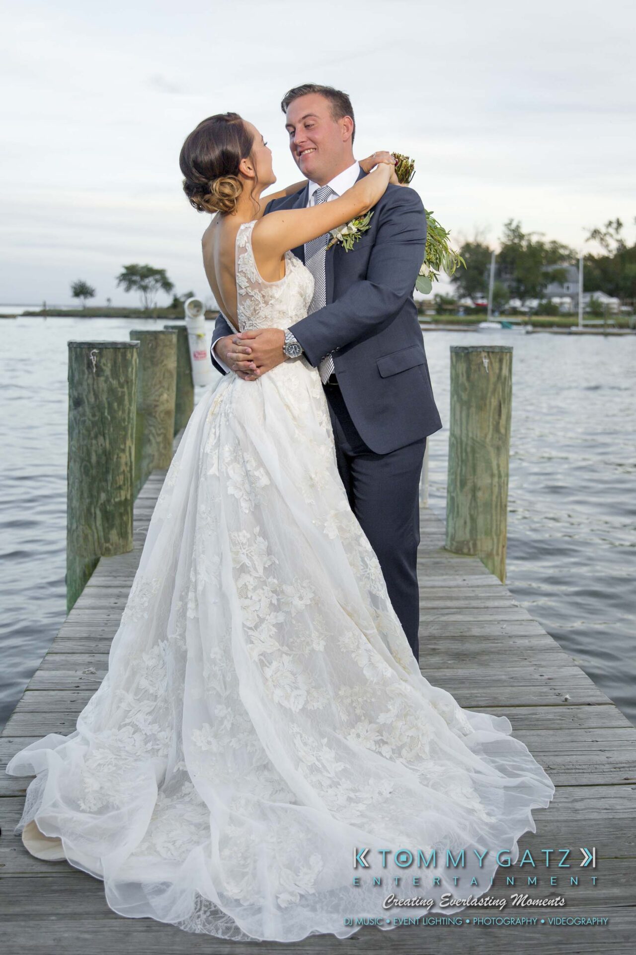 Amanda-Tyler-102117-FORMALS-Low-Res-WM-46 - TOMMY GATZ ENTERTAINMENT A couple in formal attire embraces on a dock by the water. The woman wears a white lace gown, and the man is in a dark suit. Trees and water are in the background.