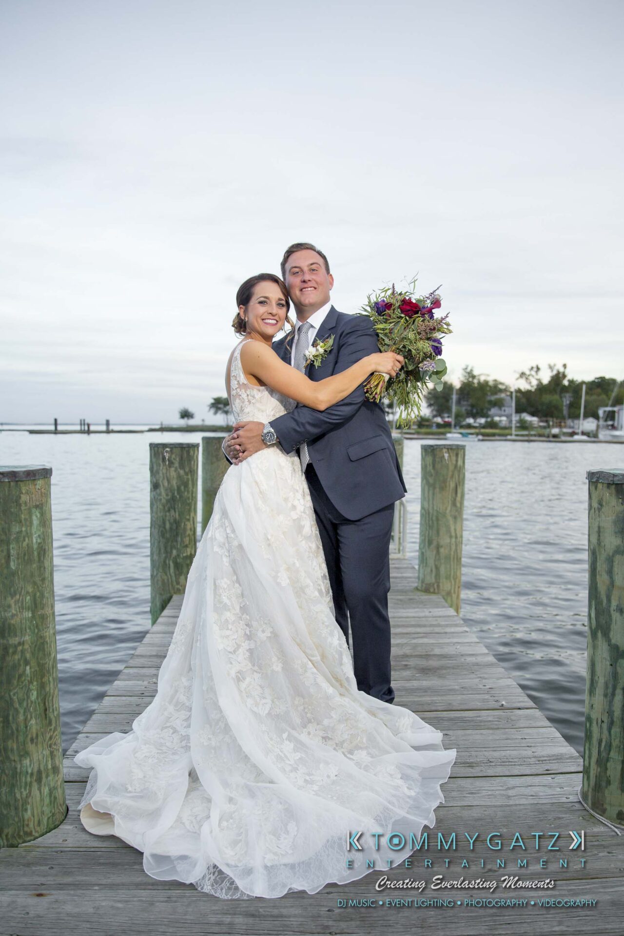 Amanda-Tyler-102117-FORMALS-Low-Res-WM-48 - TOMMY GATZ ENTERTAINMENT A couple in wedding attire embraces on a wooden dock by a body of water, with trees and a cloudy sky in the background.