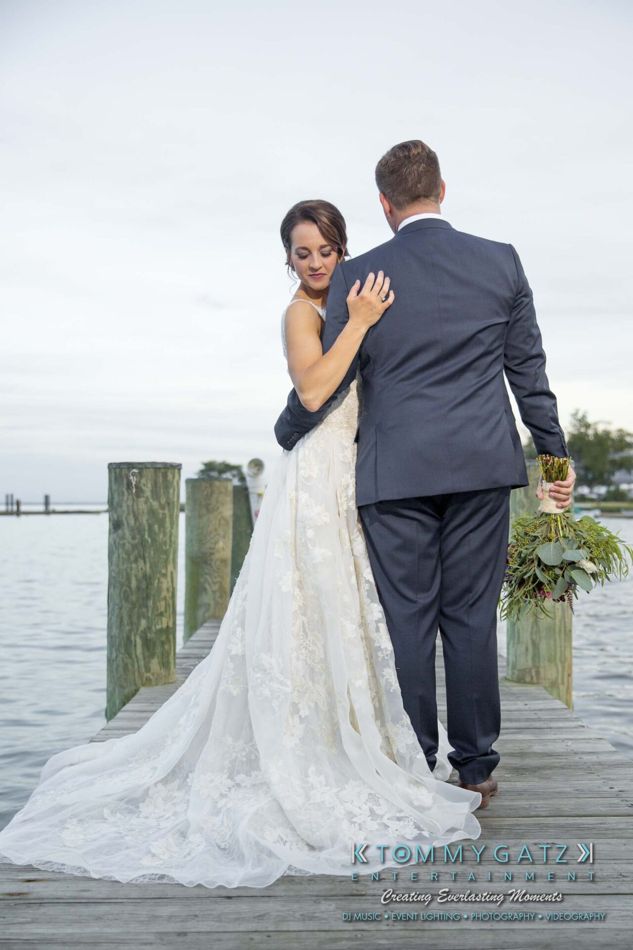 Amanda-Tyler-102117-FORMALS-Low-Res-WM-49 - TOMMY GATZ ENTERTAINMENT A bride and groom stand on a wooden dock by the water. The bride wears a white gown and holds the groom's shoulder. The groom wears a dark suit, holding a bouquet. Trees are visible in the background.